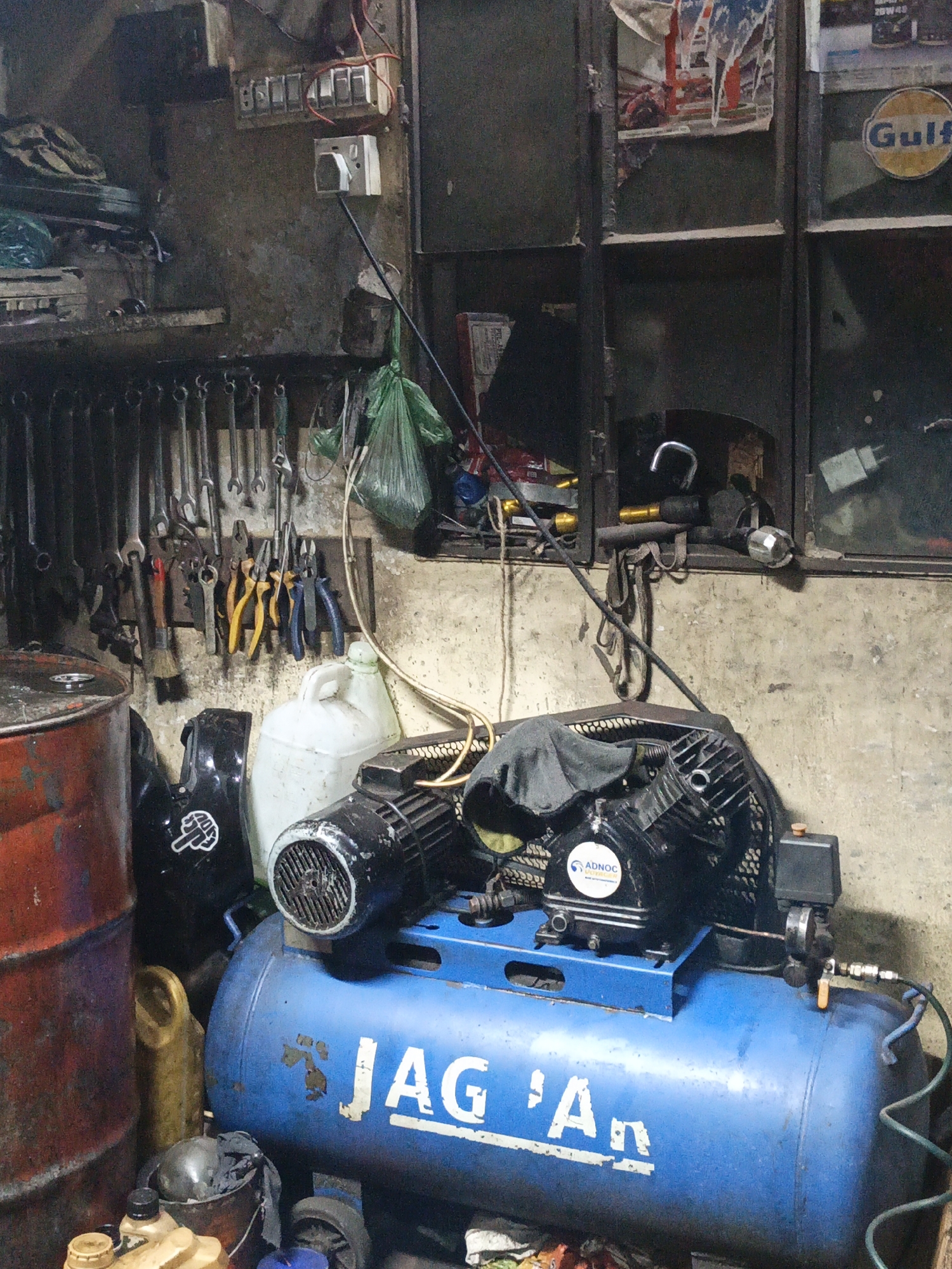 The image depicts an auto repair shop in Dhaka, Bangladesh. A blue air compressor with "JAG 'An" stenciled on it is in the foreground, situated near barrels, oil containers, and a helmet with a fist sticker. In the background, a wall displays an array of wrenches and pliers, hung above a white container. A cabinet sits above the tools, and a Gulf oil can is visible inside. Electrical outlets and wires are mounted near the ceiling. The environment is cluttered and worn, indicative of a working repair shop. No people are visible in the frame.