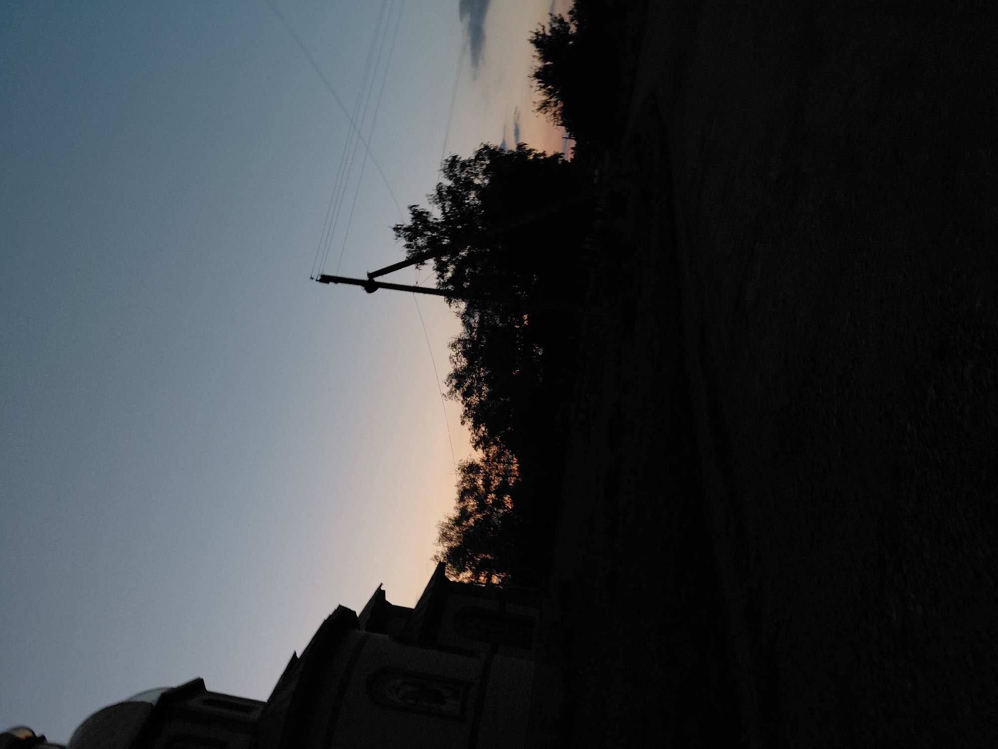 In the city of Ustyvytsia, Ukraine, a silhouette of a church stands to the left of the frame, its dome and upper sections dark against a fading twilight sky. The church building features arched windows and a mural or relief on its facade. In the mid-ground, a line of trees, also silhouetted, stretches across the horizon. A lone utility pole with overhead wires is visible amongst the trees, contributing to the rural or semi-rural setting. The foreground is dominated by darkness, obscuring the ground details, suggesting it might be a dirt road or path. The sky transitions from a pale blue at the top to a warm, peachy hue near the horizon, indicating the end of the day. The overall mood is tranquil and somewhat somber due to the low light conditions and the dark silhouettes.