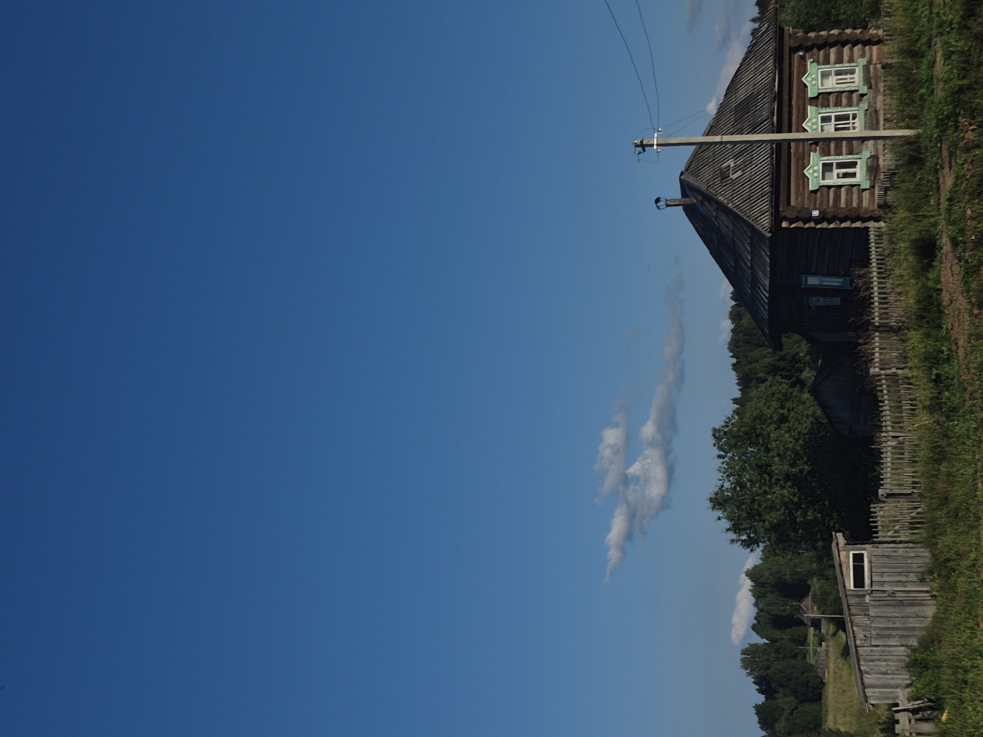 A traditional Russian wooden izba, or log house, with a dark wooden exterior and a grey, corrugated metal roof, stands on a slight incline under a vast, clear blue sky. The house features three visible windows on its front facade, each framed with intricate green wooden carvings and white sills. A simple, weathered wooden fence runs along the front of the property, with grass and vegetation growing around its base. To the left of the izba, a smaller, more rustic wooden shed with a single rectangular window is partially visible. In the background, a dense, green forest stretches across the horizon, punctuated by several trees of varying heights. A utility pole with visible wires stands to the right of the izba. The image captures a rural scene, likely in the village of Дойная, Russia, during daylight hours.