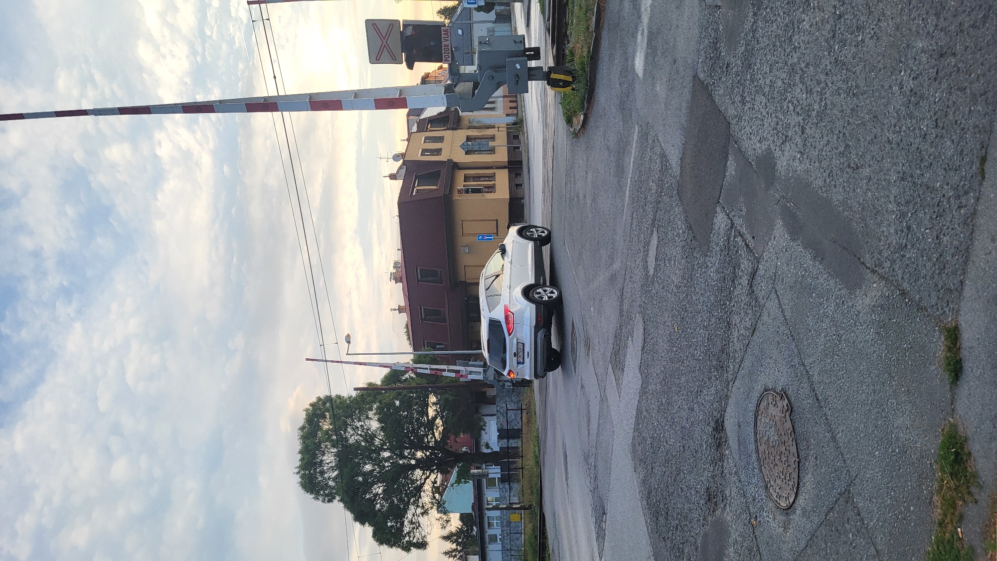 The image depicts a view of a railway crossing in Prešov, Slovakia, at dusk. A white SUV, identified by its license plate as SR-298FE, is turning at the intersection in front of the crossing. In the background, a yellow building with a red roof stands prominently, with other residential buildings and trees visible further back. A striped red and white railway barrier is raised, indicating that trains are not currently passing. A sign with the Slovak text "POZOR VLAK" (Beware of Train) is also visible. The sky is overcast with scattered clouds. The road surface is asphalt with visible patches and markings. A manhole cover is present in the foreground.