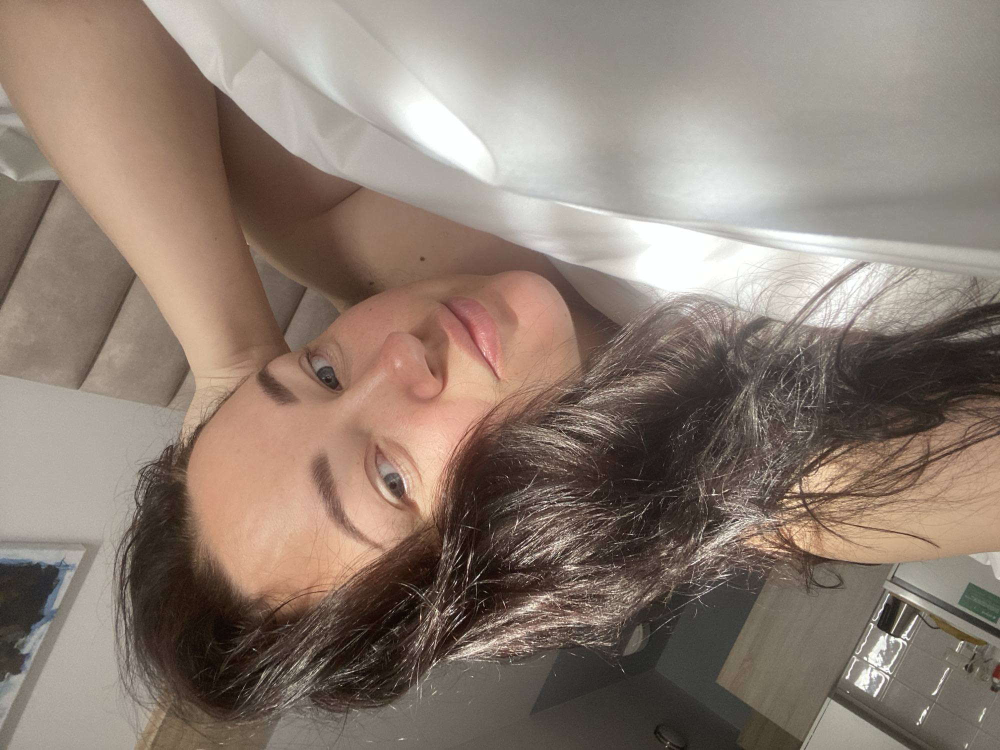 A woman with dark hair is lying down, partially covered by a white sheet. Her face is turned towards the camera. She has fair skin and blue eyes. Her left arm is positioned above her head and her right arm is near her face. The background appears to be an interior space, possibly a bedroom or living area, with a padded headboard visible on the left side. The lighting suggests daylight, with shadows present. The location information suggests this is in Sochi, Russia, though there is no evidence to corroborate that in the image.