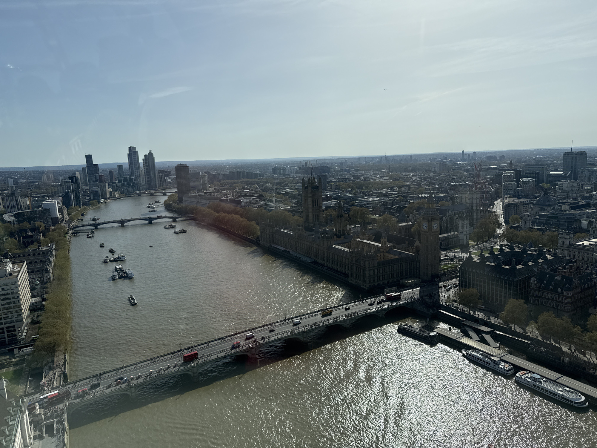 The image shows an aerial view of London, United Kingdom, featuring the River Thames, several bridges, and prominent landmarks. The Houses of Parliament and Big Ben are centrally located. Multiple bridges span the river, carrying vehicular and pedestrian traffic. Boats dot the river, and several are docked. The river reflects sunlight, creating a shimmering effect. The city's skyline is visible in the background, with numerous buildings and structures. The sky is clear and bright.