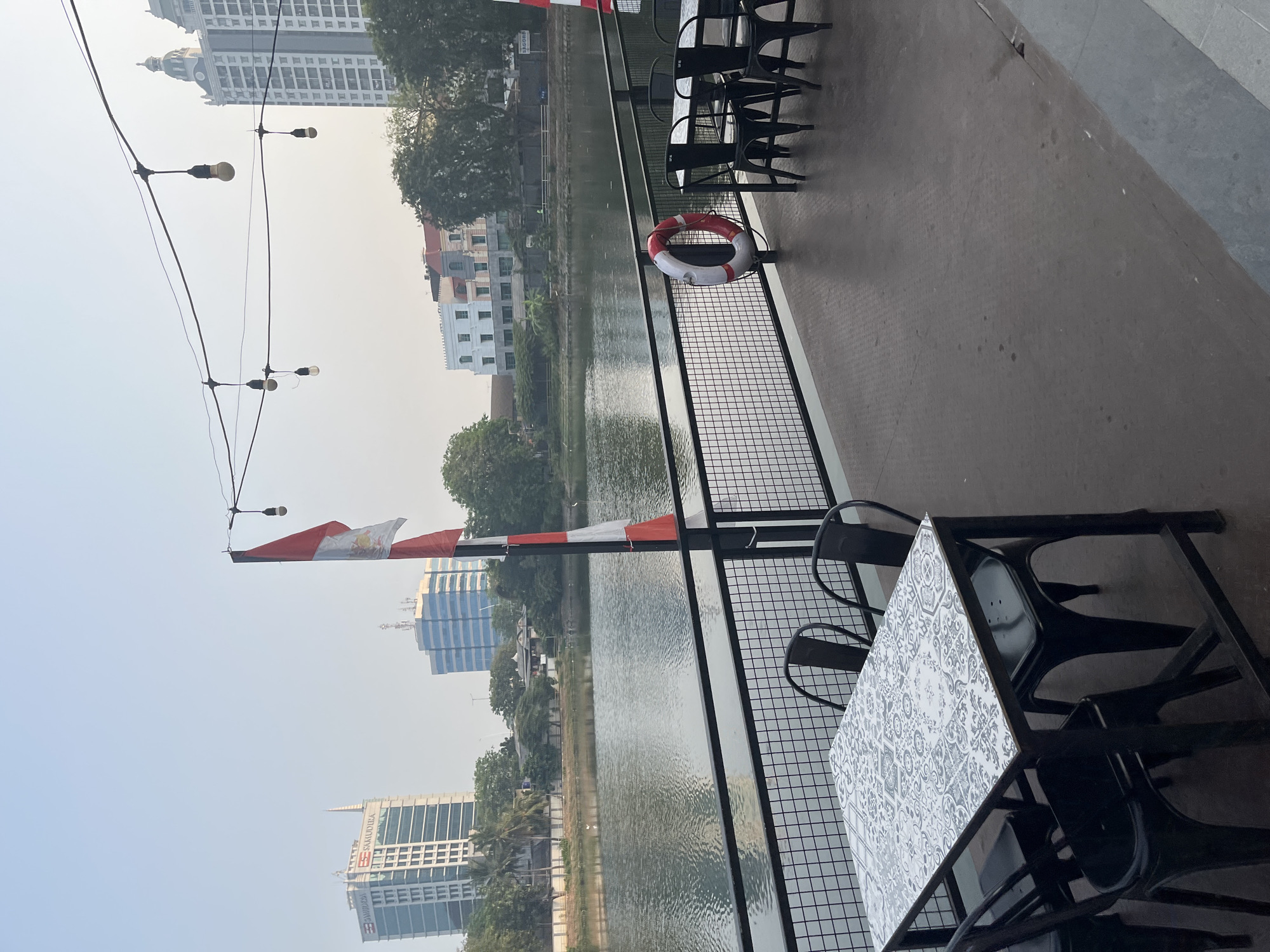 The image shows an outdoor seating area on a deck overlooking a body of water. The deck has tables and chairs, and a railing. In the background, there are several buildings and trees. A flag is visible on a pole. There are also string lights hanging overhead. A life preserver is attached to the railing.