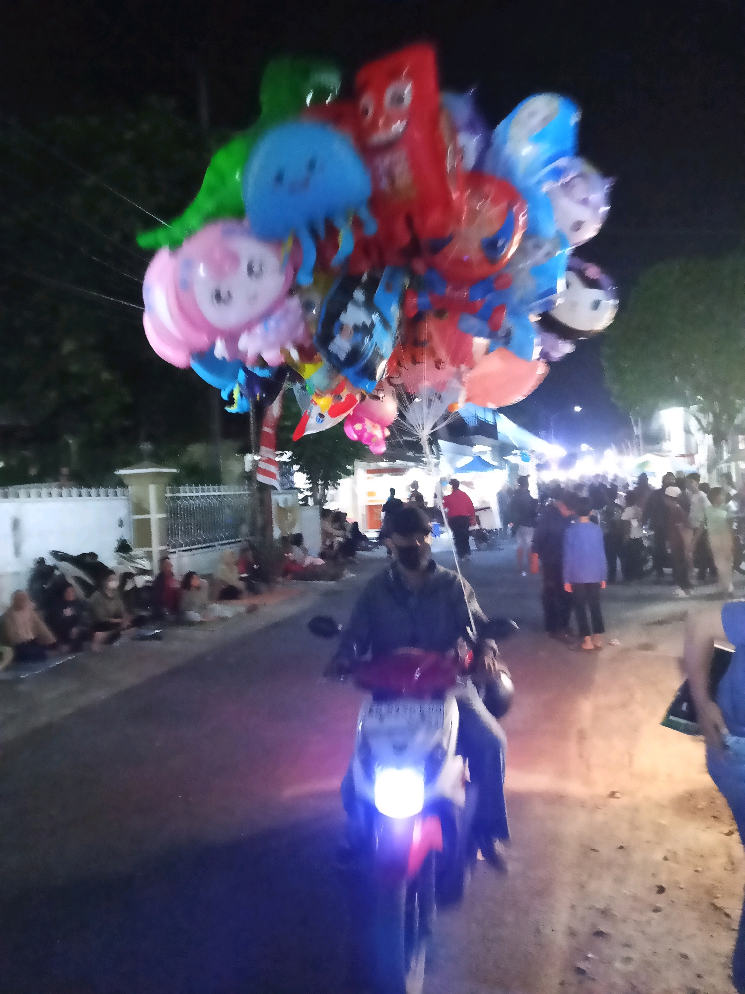 In Bendosewu, Indonesia, at night, a person on a motorcycle drives past a crowd gathered on the side of a street. The motorcycle is illuminated by its headlights. The person on the motorcycle is holding a large cluster of colorful balloons, many of which are shaped like cartoon characters and animals. The crowd consists of adults and possibly children sitting and standing, observing the scene. The street is lined with a white fence and trees. In the background, there are stalls and more people, suggesting a market or a festive event. The overall atmosphere is lively and festive, typical of a night market or local celebration.