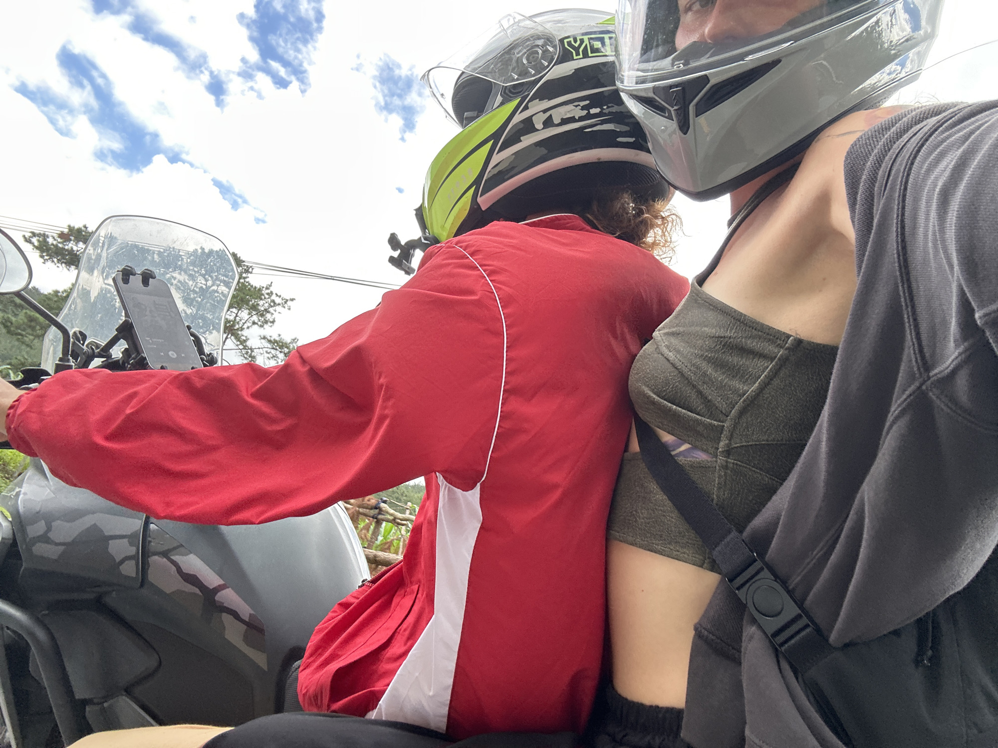 Two individuals are depicted on a dark grey motorcycle in Púng Luông, Vietnam, observed from a low, forward-looking perspective. The sky, partially blue with elongated white clouds, occupies the upper portion of the image.
The individual in the immediate foreground, positioned on the right, wears a light grey motorcycle helmet with a clear visor, revealing the right eye and part of the nose. This person is dressed in a dark olive green strapless top and has a dark grey hooded garment draped over their left shoulder, secured by a black strap with a central buckle. A tattoo is visible on their left shoulder.
Behind and to the left of the foreground individual, another person, likely the driver or front passenger, wears a red jacket featuring a vertical white stripe on its left rear side. This individual's helmet is grey with a black, white, and neon green pattern, displaying the "YETI" brand name in green. Curly brown hair is visible protruding from the base of this helmet. The right arm, covered by the red jacket, extends forward towards the motorcycle's handlebars.
The motorcycle is dark grey and equipped with a clear windscreen. A mobile phone, displaying an application interface, is mounted on the handlebars, visible through the windscreen. The motorcycle's side panel exhibits a subtle grey camouflage pattern, and a black protective bar is partially visible on the left. In the background, green foliage and thin black utility lines are present.