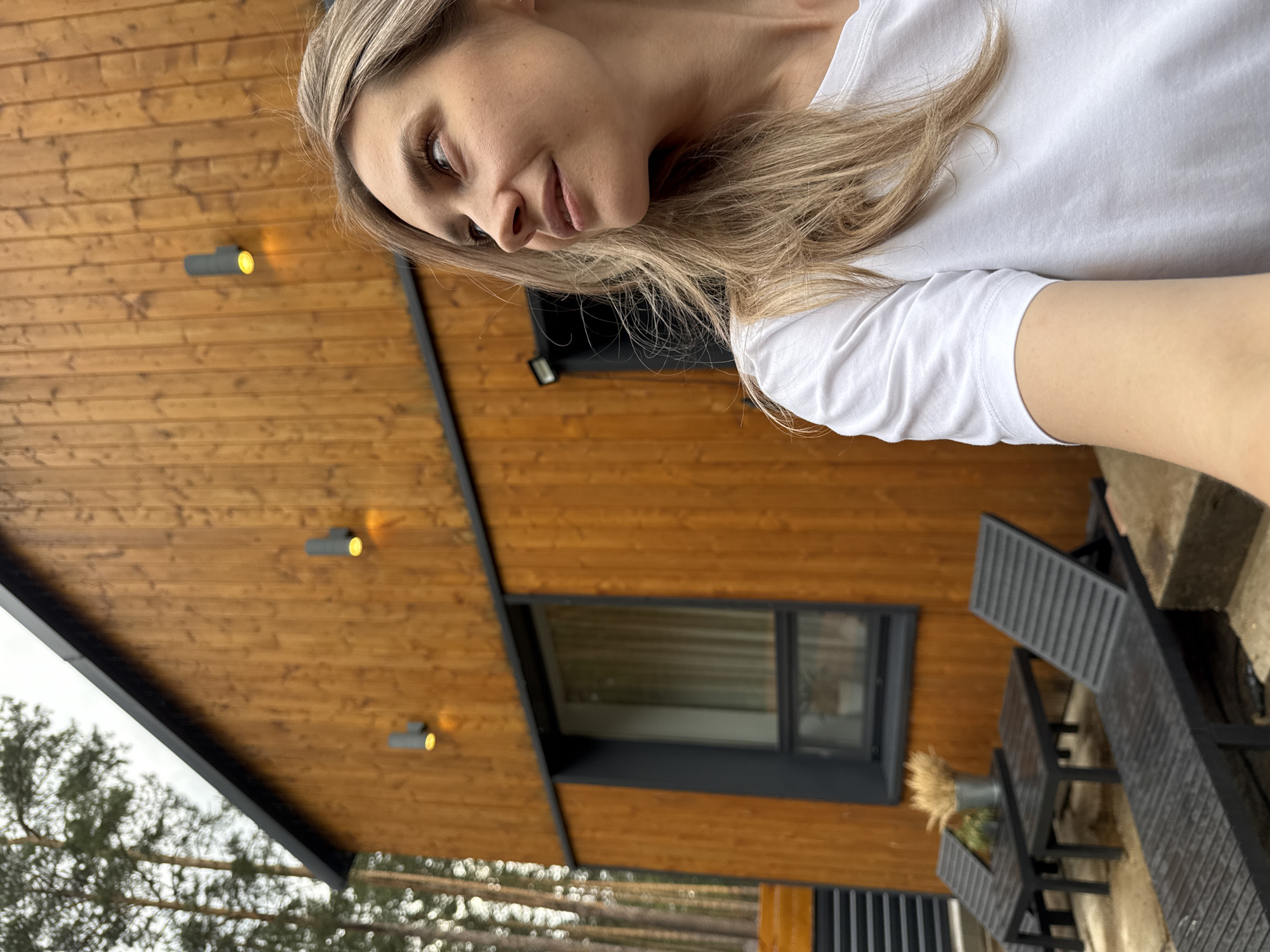 A woman with blonde hair is taking a selfie in front of a modern wood-paneled house. The house has black trim and multiple outdoor lights. In the background, there are trees and potentially other parts of the house's outdoor area. The location is possibly in Chaykovsky, Russia.
