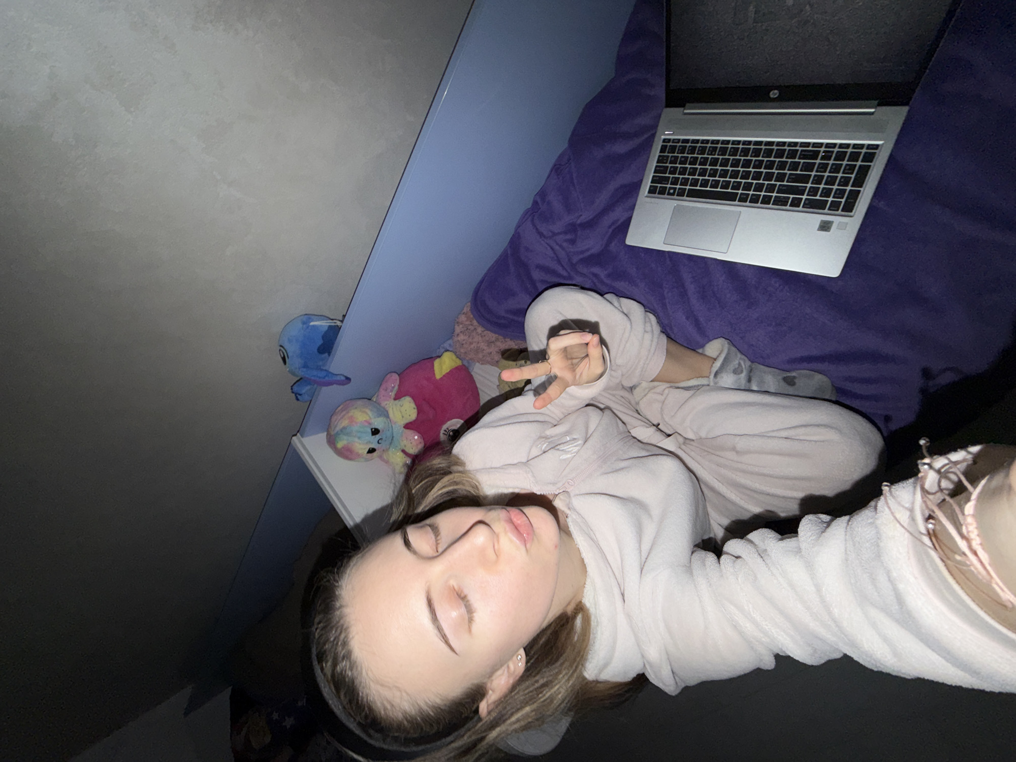 A young woman is lying on a bed with a purple blanket. A laptop rests on the blanket. Several stuffed animals are visible next to her, including a blue Stitch plushie and a pink plushie. The room is lit by dim lighting. The location appears to be an indoor residential space, possibly a bedroom. The location in Plovdiv, Bulgaria cannot be determined from this image.