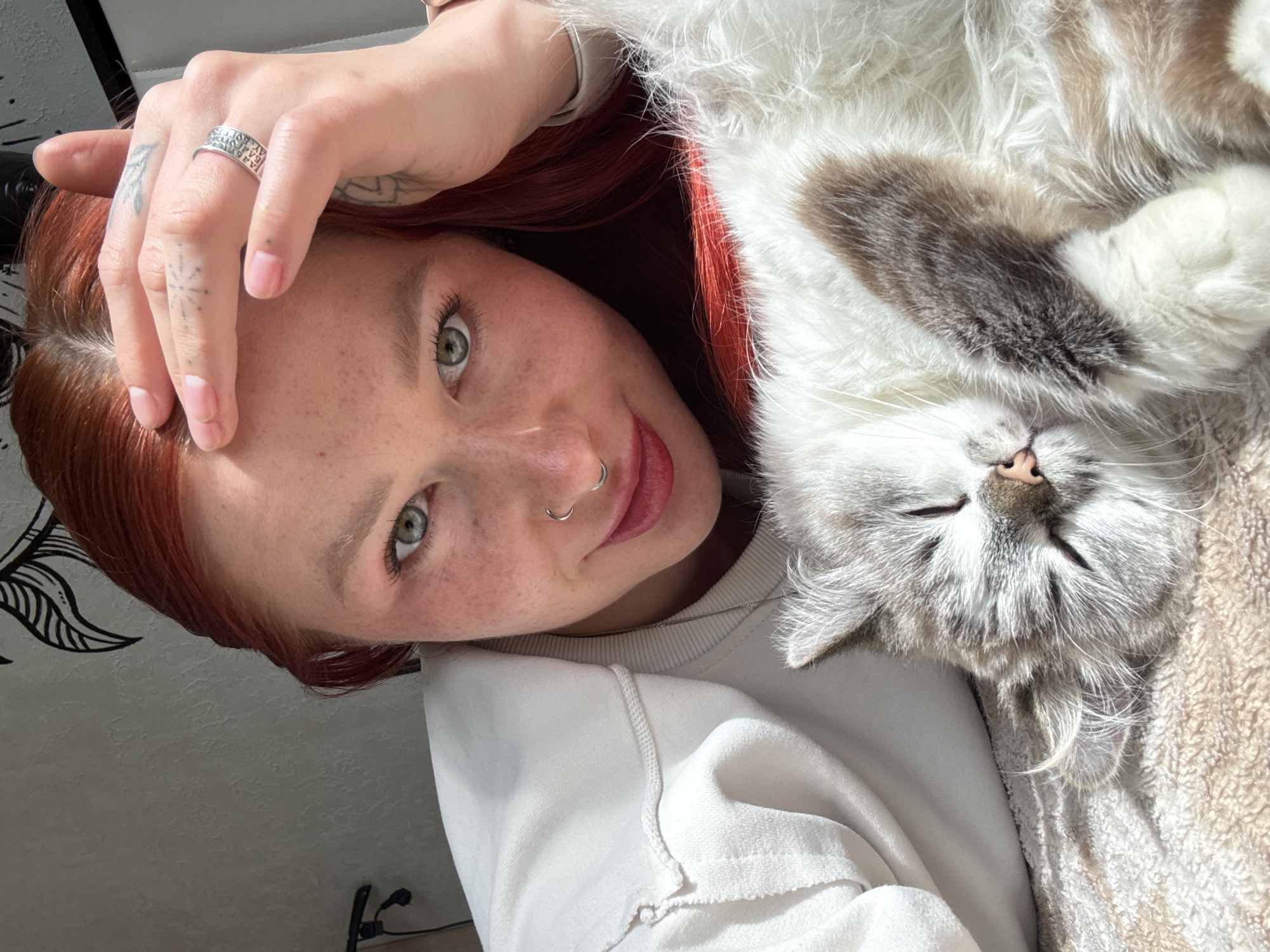A high-angle, close-up photograph captures a young woman and a cat in a relaxed indoor setting, likely in Rostov-on-Don, Russia. The woman, positioned on the left, has striking red hair, fair skin with noticeable freckles, and vibrant green eyes. She is smiling gently towards the camera. She sports a silver septum piercing and a small lip ring. On her right hand, which is raised towards her head, she wears a silver ring with an intricate patterned design that includes repeated symbols or letters. Small, faded tattoos, including a compass rose on her middle finger, are also visible on her hand. She is dressed in a light-colored (possibly white or off-white) sweatshirt or sweater.
Next to her, on the right, a fluffy, long-haired cat rests upside down, appearing to be deeply asleep or very relaxed, with its eyes closed. The cat's fur is a mix of white, light grey, and brown patches, particularly prominent on its chest and paws. Its small pink nose is visible. Both subjects are illuminated by bright natural light, indicating a daytime scene. The cat is lying on a soft, textured beige or light brown blanket. The background shows a light-colored wall adorned with a partial black and white decorative drawing, resembling a plant or feather motif. The overall atmosphere conveyed is one of warmth, affection, and peaceful companionship.