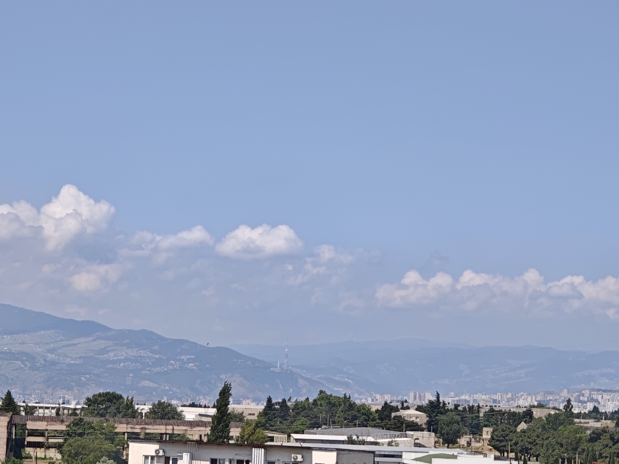 The image is an outdoor daytime shot of a city skyline and distant mountains under a clear blue sky with scattered white clouds. In the foreground, there are several buildings with flat roofs and visible air conditioning units. A few evergreen trees are scattered around the buildings. The middle ground shows a dense line of trees and more buildings, some of which appear to be industrial or commercial. Further in the distance, rolling hills and mountains are visible, with a city spread across the lower slopes and a television tower on one of the peaks. The farthest background shows a hazy mountain range. There are no people or animals visible in the frame.