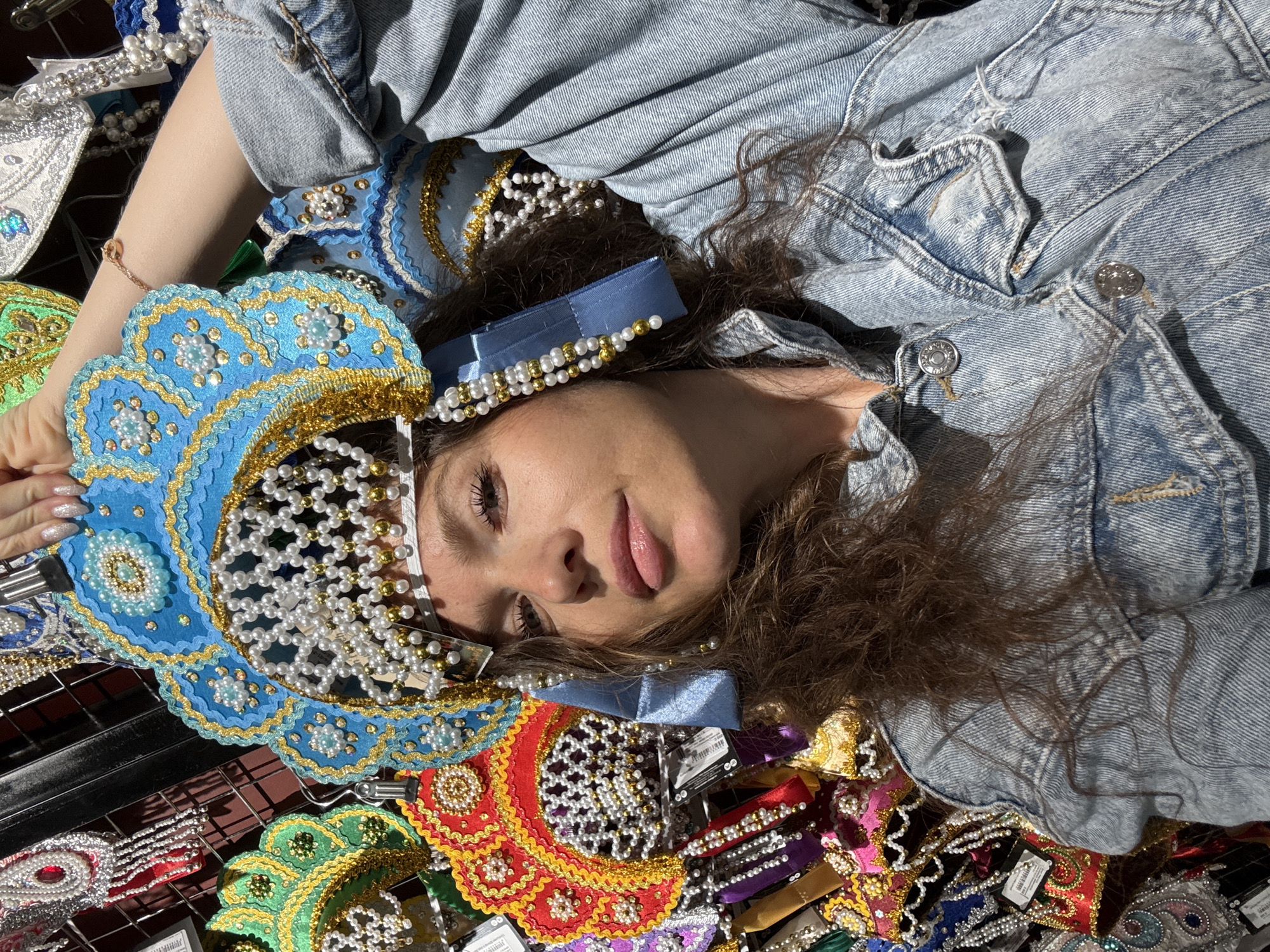 A young Caucasian woman with long, wavy brown hair is photographed from the chest up in a selfie. She is wearing a light-wash denim jacket. On her head, she wears a large, ornate blue and gold headdress adorned with numerous white pearls and gold beads. The headdress has a circular base and intricate, fan-like extensions. She is holding the headdress up with her right hand, her arm extended. Her eyes are open and she is looking directly at the camera, her lips slightly parted. The background is filled with a display of similar colorful, bejeweled headdresses and headwear, suggesting she is in a market or store. The lighting is bright and appears to be natural sunlight, casting shadows and highlighting the textures of the clothing and headdresses. The location is not specified beyond "Unknown, Unknown."