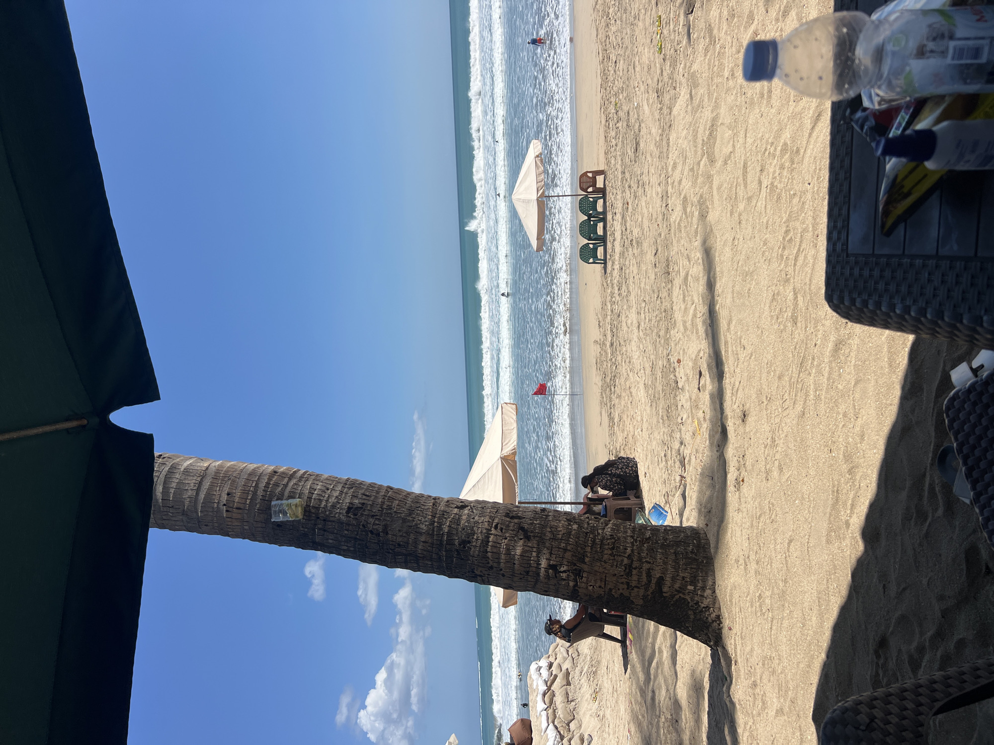 This image depicts a sunny day at Kuta Beach, Indonesia, characterized by a sandy shoreline and clear blue skies. In the foreground, a portion of a large umbrella shades a woven table with a plastic bottle of water and other items. A prominent palm tree stands to the left, with a glass attached to its trunk.
Further back, several beach umbrellas are set up on the sand, with chairs placed beneath some of them. Two people are seated under one umbrella near the palm tree, and a red flag is visible on a pole in the distance. The ocean features moderate waves, with several surfers in the water. A person in red shorts is wading in the shallow water to the right. In the background, the coastline is lined with sandbags. The overall scene captures a typical day at a popular beach destination.
