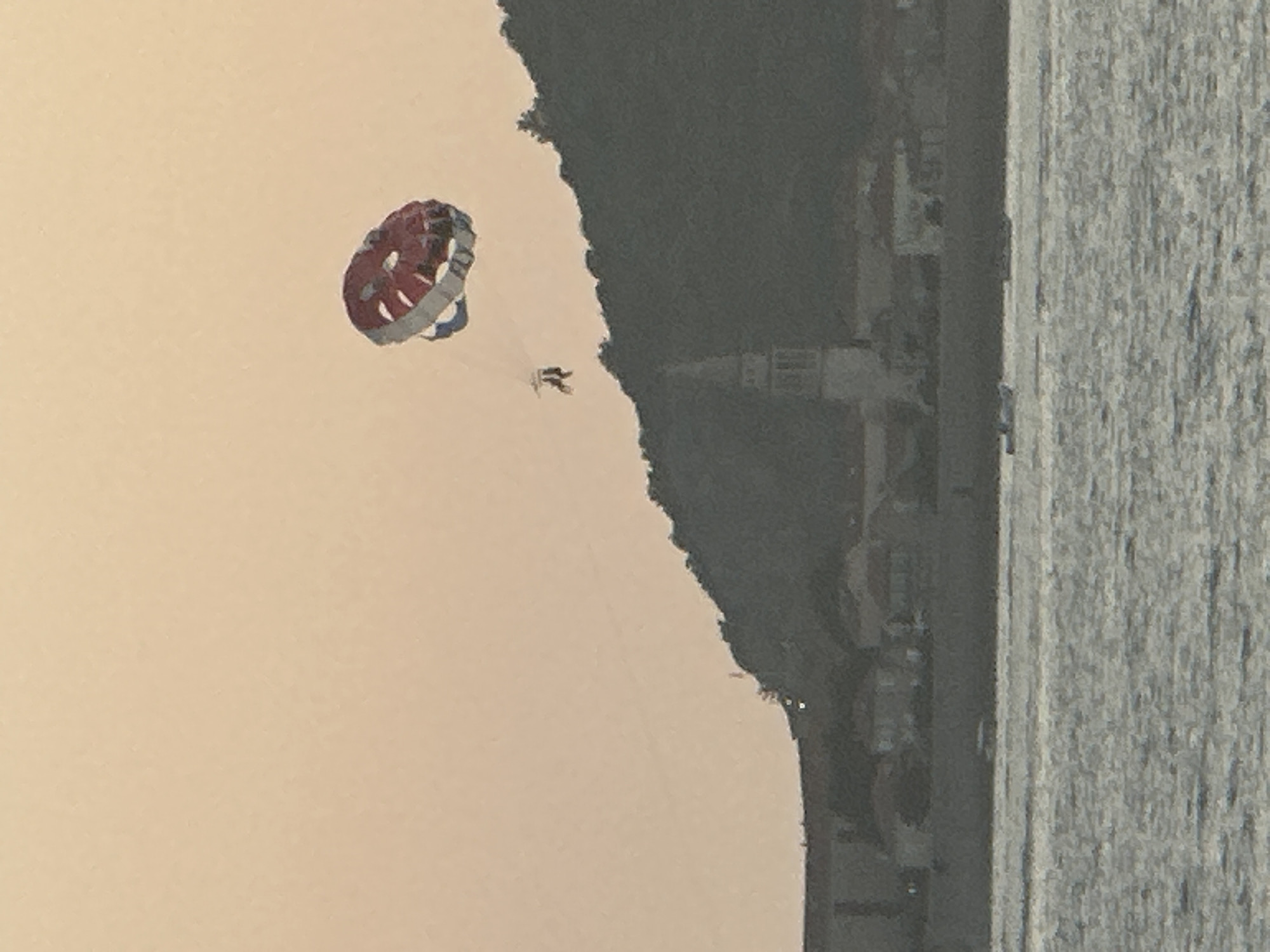 The image shows a parasailer being towed over the water in front of Budva, Montenegro. The parasail is red and white with blue trim. Two people are visible in the harness below the parasail. In the background, a tree-covered hill rises behind a town with buildings and a prominent bell tower. The water is choppy, and a few small boats are visible on the surface. The sky is a hazy orange, suggesting either dawn or dusk.