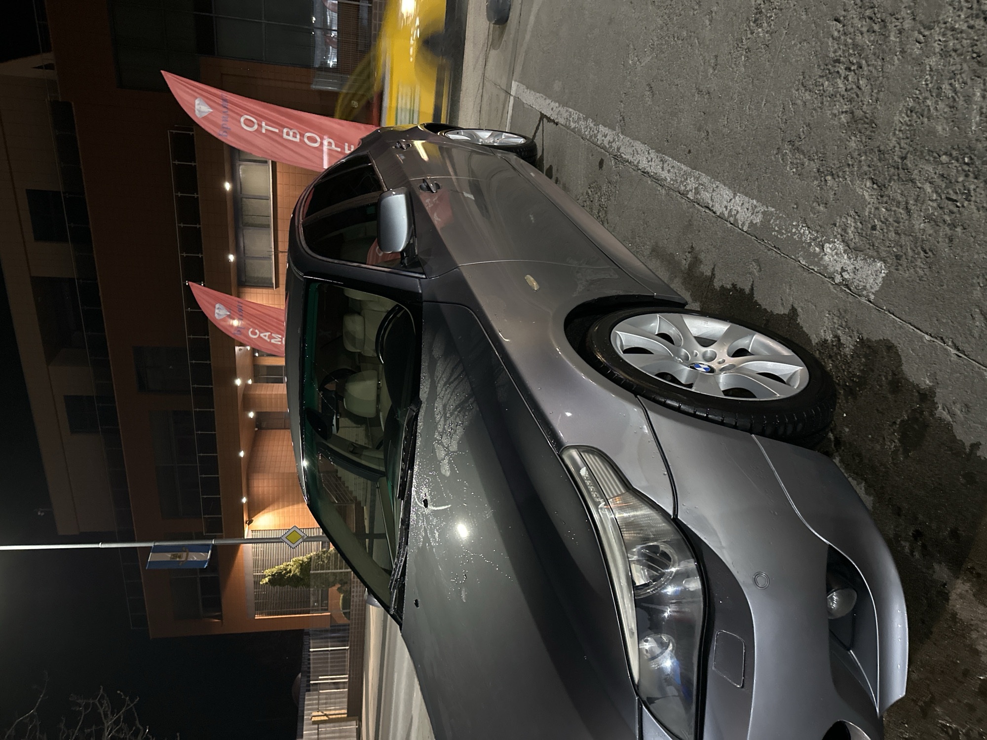 A grey BMW 5-series sedan is parked on asphalt in front of a building in Plovdiv, Bulgaria, at night. The building has red banners with white text in front, as well as a flag and street sign. The car is clean but has some water droplets on its surface. It is parked next to a white parking line on the right. The sky is dark, and there are trees visible in the background.
