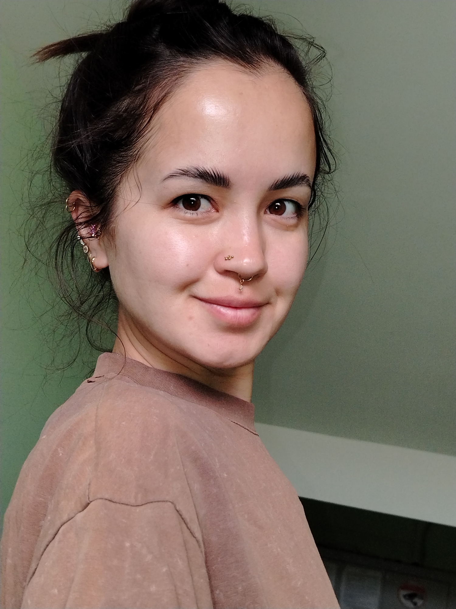 A close-up shot features a woman with dark hair styled in a messy bun. She has fair skin and brown eyes, and a subtle smile. She has multiple gold-toned piercings in her ear, including hoops and a floral stud. Additional gold-toned piercings are visible in her nose, including a stud on the nostril and a septum ring. She's wearing a brown t-shirt. The background is a muted green color. The location in the city of Noviy, Russia is not apparent from the given image.