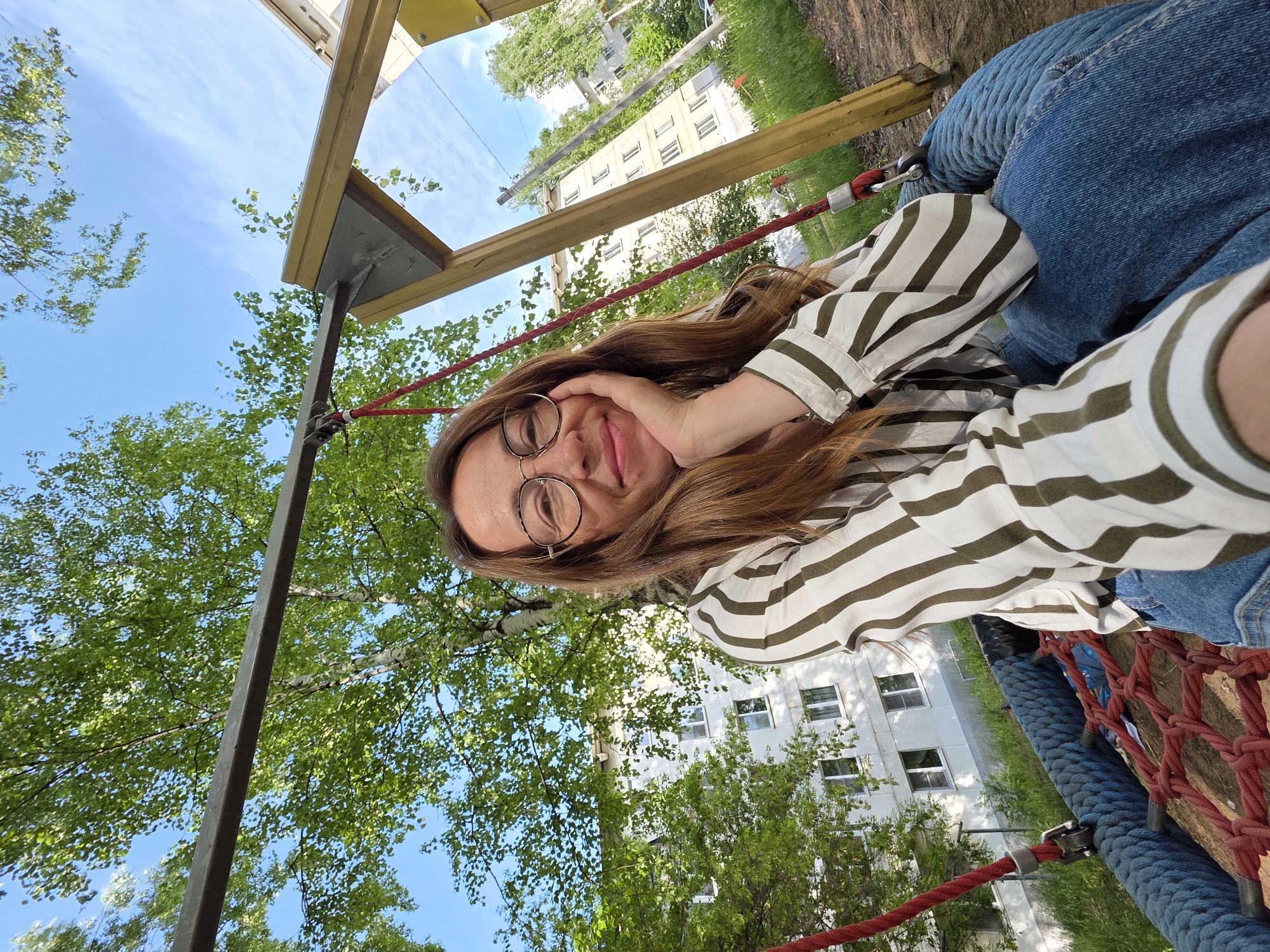 A young woman with long brown hair and glasses is in a playground in Chaikovsky, Russia, taking a selfie. She is wearing a striped shirt and blue jeans. She is sitting on a blue rope net, and a red rope is visible. Behind her are trees and a white building with windows. A yellow structure with a metal support is also visible. The sky is blue with some clouds.