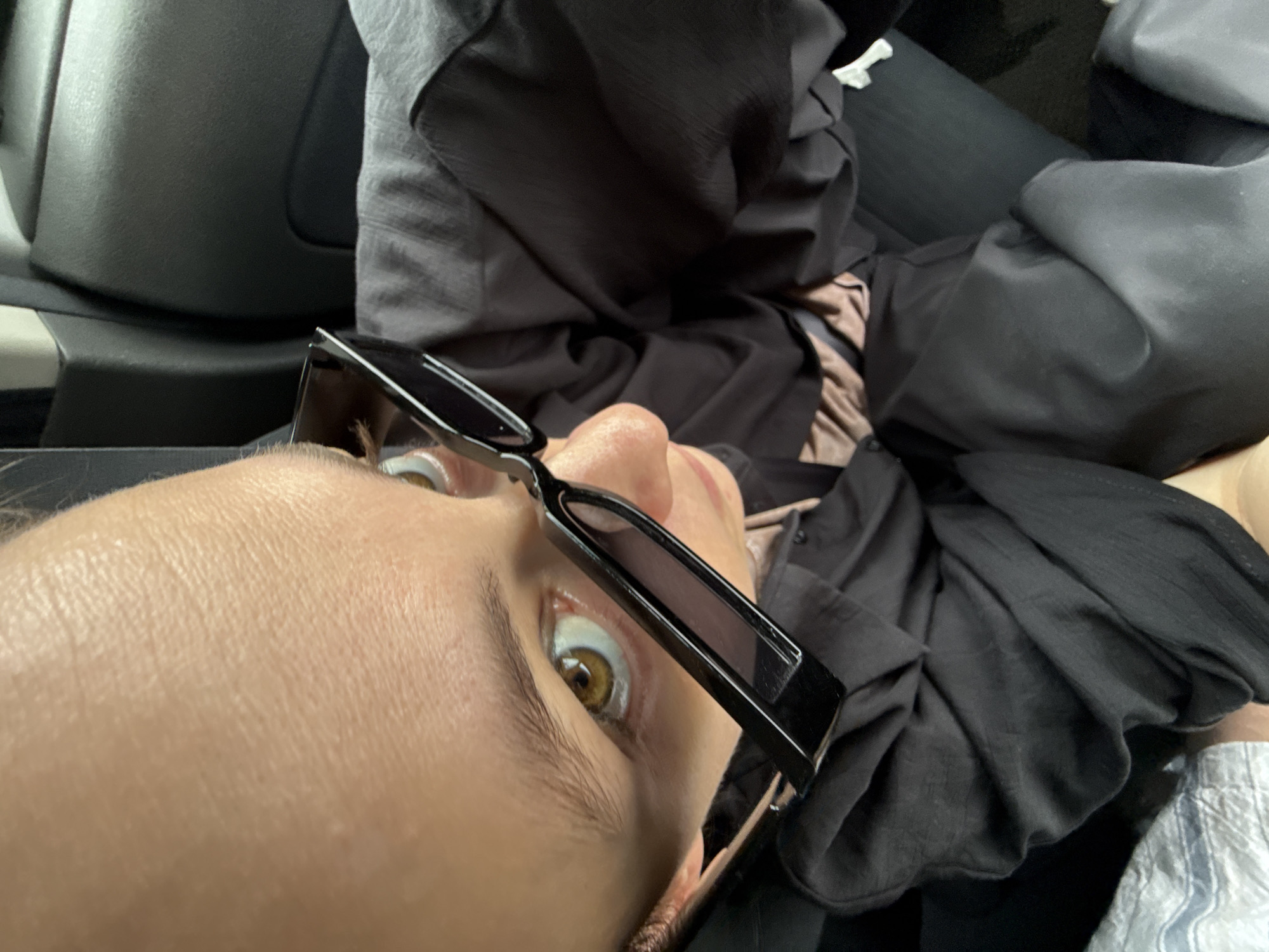 A close-up, low-angle shot shows a person lying down, possibly in a car. The person is wearing sunglasses, which are partially obscuring their face, but one eye is visible, revealing a brown iris. They are wearing a black garment, and there is a grey car seat visible. The environment appears to be inside a vehicle. Kukeevo, Russia, is not identifiable from the image.