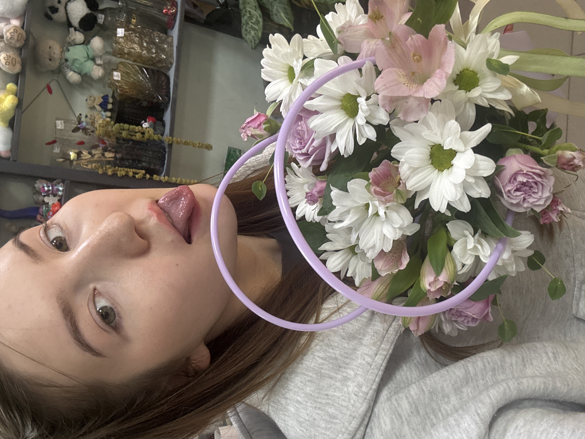 A medium shot shows a young woman in a grey hooded sweatshirt holding a bouquet of flowers in a basket with purple handles. She is leaning into the frame, sticking her tongue out slightly. The bouquet consists of white daisies, pink alstroemeria, and a few roses, some of which are budding. In the background, shelves are visible displaying various items, including stuffed animals and decorative vases, suggesting the location is a shop. The overall lighting is soft.