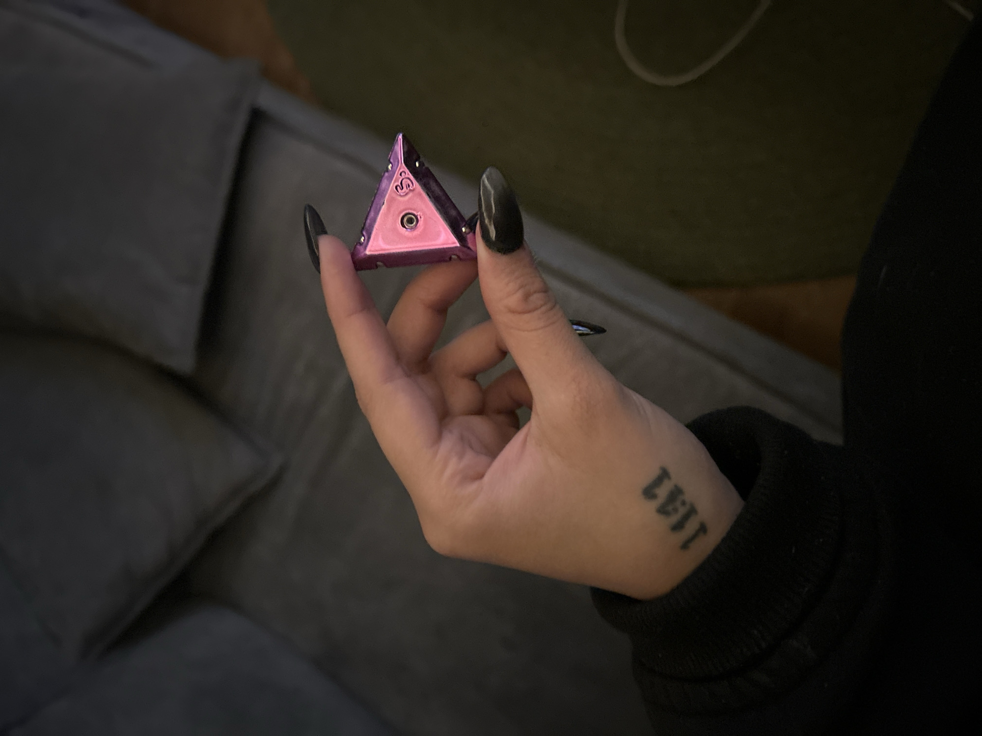 A person is holding a pink, triangular object in their right hand. The object appears to be a metal container of some sort. The person has long, black fingernails and a tattoo on their left wrist. The background includes a grey sofa and an off-white surface. The environment cannot be attributed to Huixquilucan de Degollado, Mexico as there are no identifying characteristics.