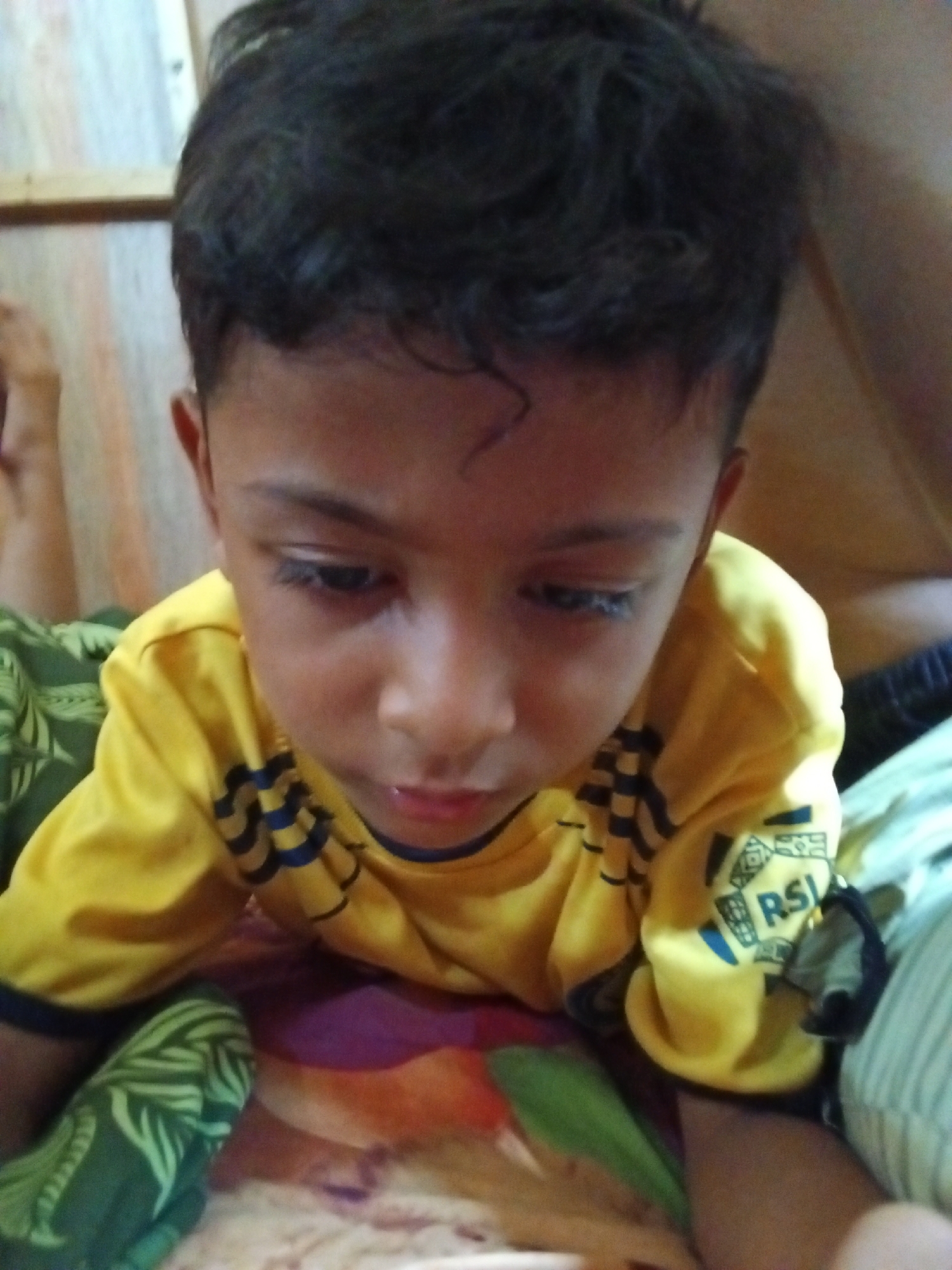 A medium shot captures a young boy from the chest up, wearing a yellow t-shirt with dark blue accents. The boy is looking down and to his right, with his head slightly tilted. His dark hair is styled with a side part, and a few strands curl over his forehead. He has dark eyes and full lips. The t-shirt has a dark blue collar and a pattern of dark blue stripes across the chest. On the right sleeve, a logo with the letters "RSI" is visible. The background is blurred, but appears to be an indoor setting with a wooden wall on the left and some striped fabric on the right. The boy is lying on a surface covered with green and patterned fabric, possibly a bed or blanket. The image is underexposed, with a reddish cast, and is grainy. The overall scene suggests a casual, personal moment.