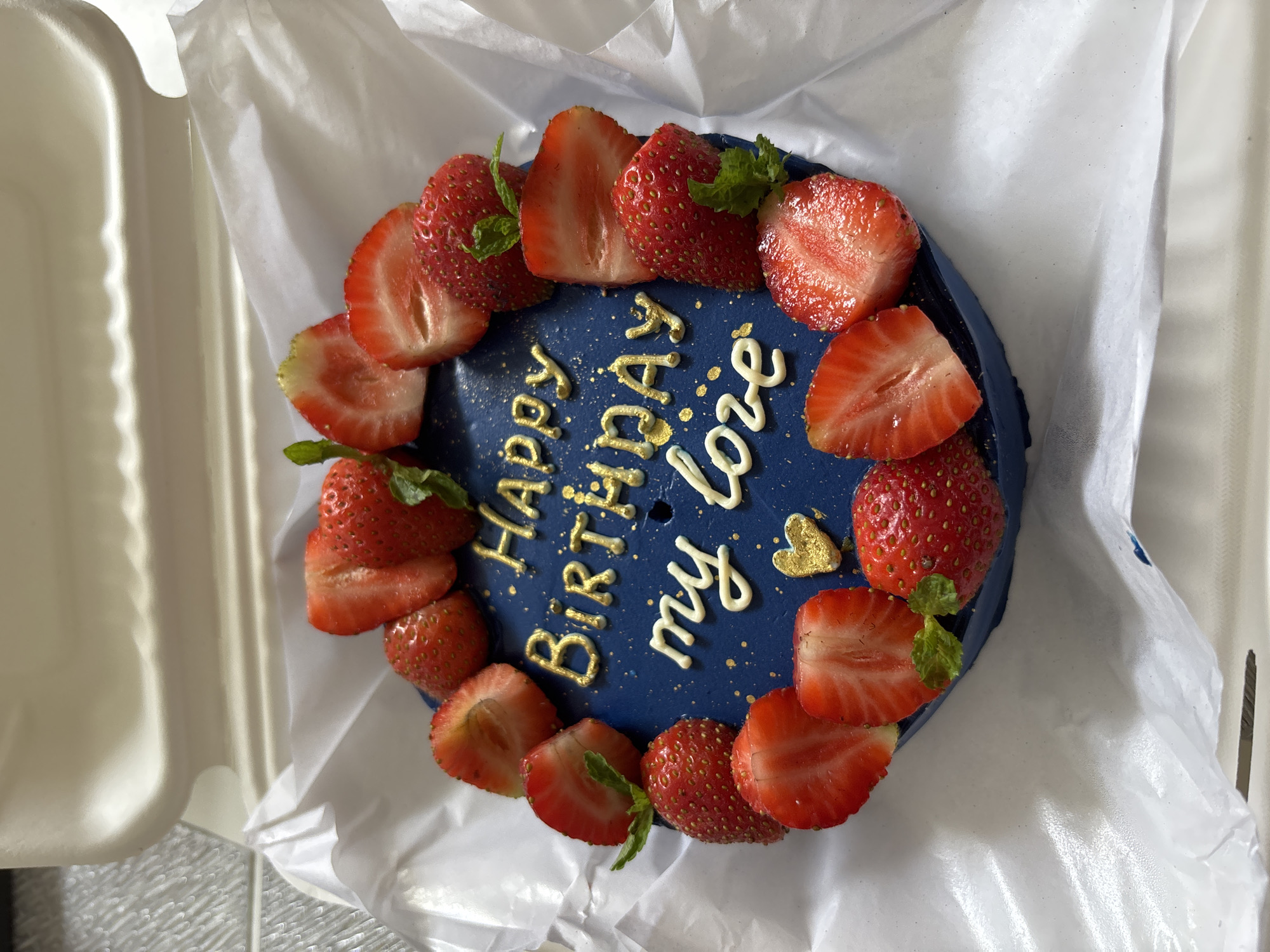 A top-down view shows a round blue cake decorated with halved and whole strawberries. The cake is adorned with golden lettering that reads "Happy Birthday my love", and a gold heart. The cake is presented in a white box on a tile surface. The location is Kuta Selatan, Indonesia.
