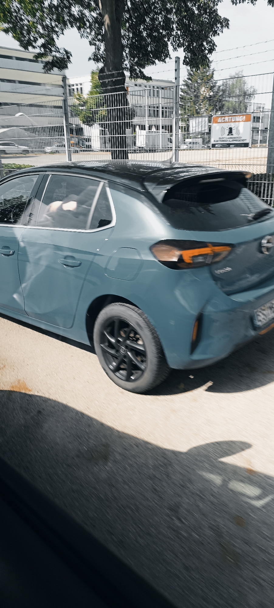 The image shows the rear passenger side of a blue Opel Corsa hatchback driving past a fence. The car has a black roof and black alloy wheels. The car's rear lights are visible, with amber turn signals illuminated. Inside the car, two people are visible in the rear seats, appearing to be looking towards the driver.
Behind the car and fence, a white building with multiple windows is visible. A sign with "ACHTUNG!" and a siren symbol is affixed to the fence. The ground is asphalt.
The image was taken in Fellbach, Germany, specifically near the border with Stuttgart, along the Bundesstraße 14. The time of day appears to be daytime, with sunlight casting shadows on the ground.