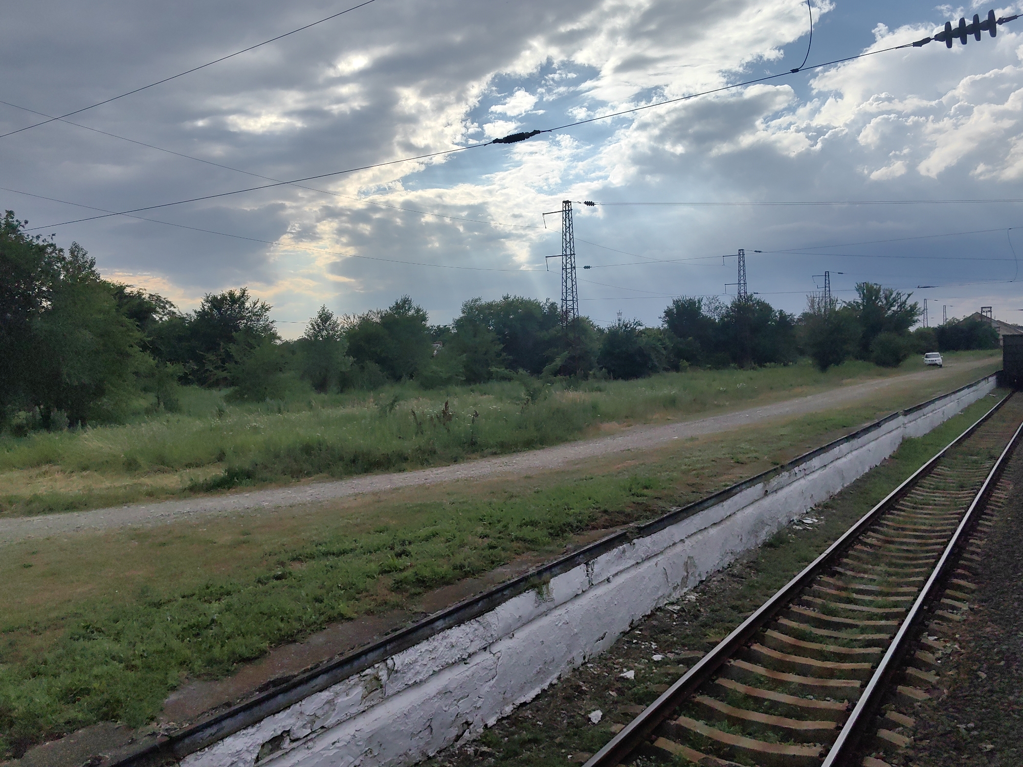 The image shows a view of a railway track and its surroundings in Novokubansk, Russia. In the foreground, a train track runs diagonally from the lower right to the upper left of the frame. The track is bordered by a low, white concrete barrier, which shows signs of wear and peeling paint. Beyond the track, a dirt path runs parallel to it, separating the track area from a field of green grass and vegetation. In the middle ground, a line of trees and bushes forms a dense green barrier. In the background, several tall electrical pylons with overhead wires are visible, indicating the presence of a power transmission line. The sky is partly cloudy with patches of blue sky and sunlight breaking through the clouds. A white vehicle is visible in the distance on the dirt path, near the trees. The scene appears to be a rural or semi-rural area within the city, likely along the outskirts where infrastructure like railways and power lines are common.