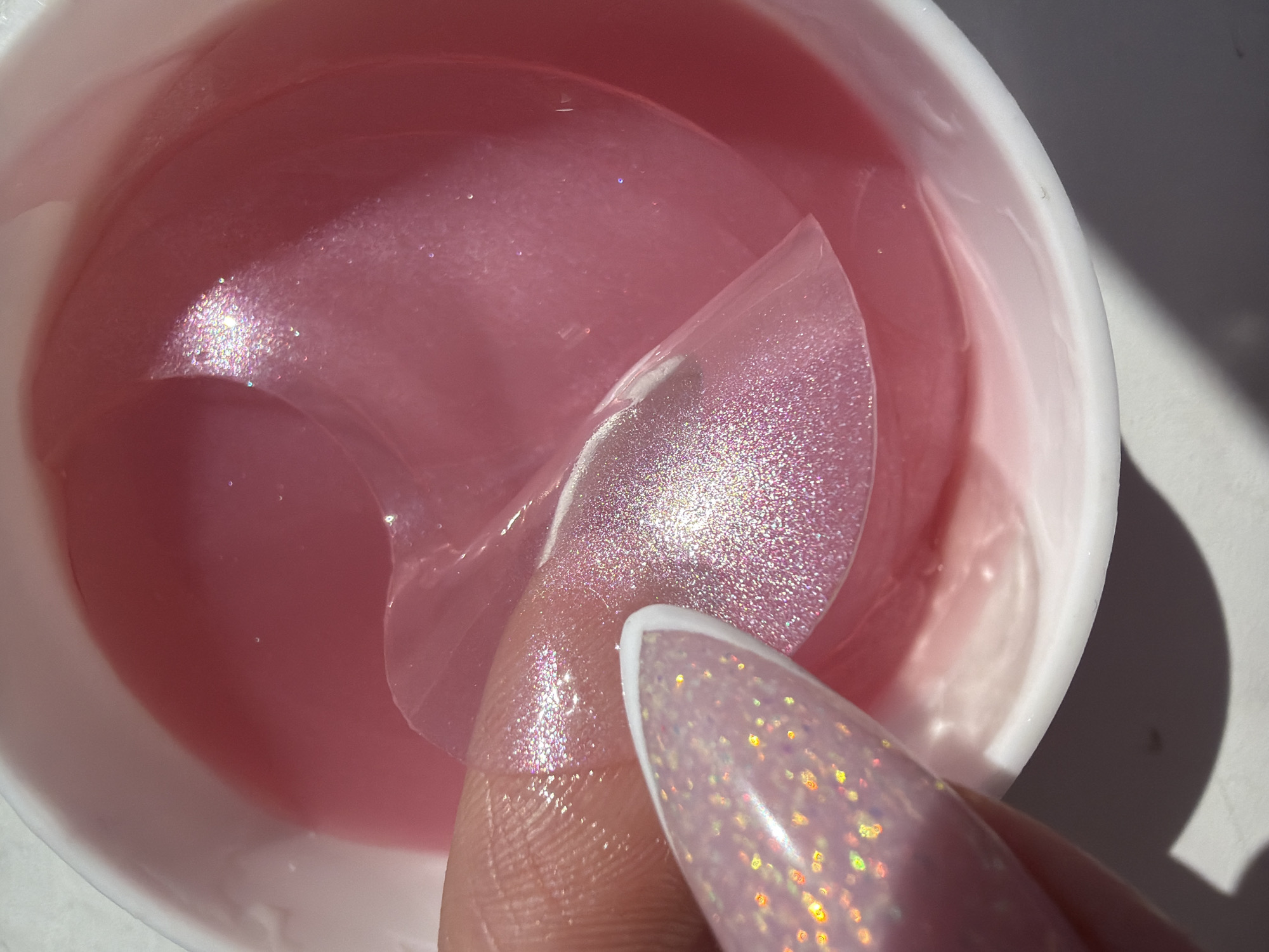 A close-up shot shows a container of pink, iridescent eye patches being held by a hand with a long, pointed nail. The nail is white with a glittery, iridescent design. The container appears to be white plastic and is filled with a pink, gel-like substance in which the eye patches are submerged. A single eye patch is being lifted from the container. The background is blurry and does not contain sufficient information to definitively identify it as Saint Petersburg, Russia.