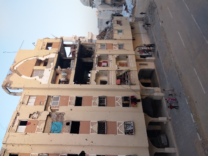The image displays a multi-story residential building in Khan Yunis, Palestinian Territory, exhibiting severe damage, likely from shelling or an explosion. The building's light-colored (beige or light yellow) facade is extensively compromised, with large sections of walls and floors completely destroyed, exposing interior rooms, collapsed concrete slabs, and twisted rebar. The central part of the structure shows the most catastrophic damage, with multiple floors appearing charred and entirely collapsed.
Despite the widespread destruction, elements of daily life are evident. Clothes are visible hanging to dry on balconies and from damaged window openings on various floors, suggesting continued habitation or recent activity. A paved road runs alongside the building. On the sidewalk, several individuals are present; a group of three or four people, including children, are seen in the lower right foreground. Further along the road, near a red and white truck, a few more figures are discernible. A white car is parked beneath an arched section of the building's ground floor. In the background, a large white-domed structure, possibly a mosque, is partially visible.
The scene, illuminated by soft, clear light suggesting either early morning or late afternoon, powerfully conveys devastation and hardship in a conflict zone, while also hinting at human resilience. No discernible text is visible in the image.