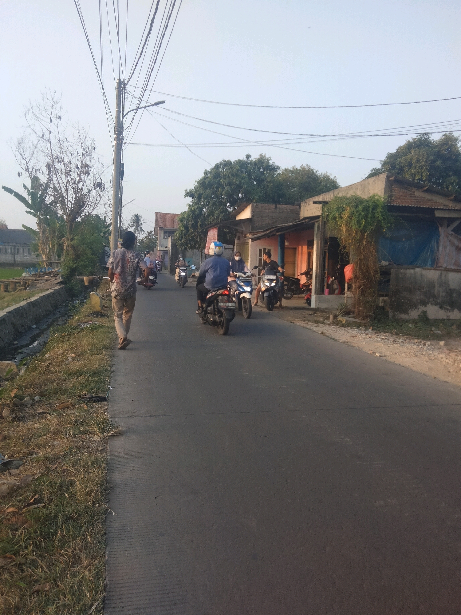 In Suka Sari, Indonesia, a person walks away from the camera on a paved road, their back to the viewer. They are wearing a patterned shirt and light-colored pants. The road is lined with a grassy verge and a drainage ditch on the left. Several motorcycles are seen on the road, some with riders. Buildings, including what appear to be shops and houses, line the right side of the road. Overhead, numerous electrical wires are strung between poles. The sky is clear and pale.
