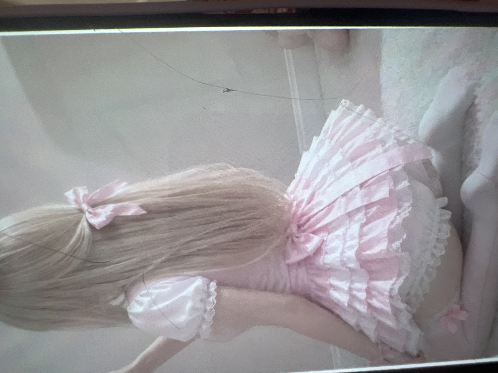 The image shows a person from behind, wearing a pink, frilly dress with white lace trim. The person has long blonde hair tied back with a pink bow. They are kneeling on a soft, light-colored surface, possibly a rug. The background is a plain, light-colored wall. The image appears to be a digital photograph, with some visible scratches or marks on the surface it is displayed on. There is no indication of the city of Kara-Dıykan, Kyrgyzstan.