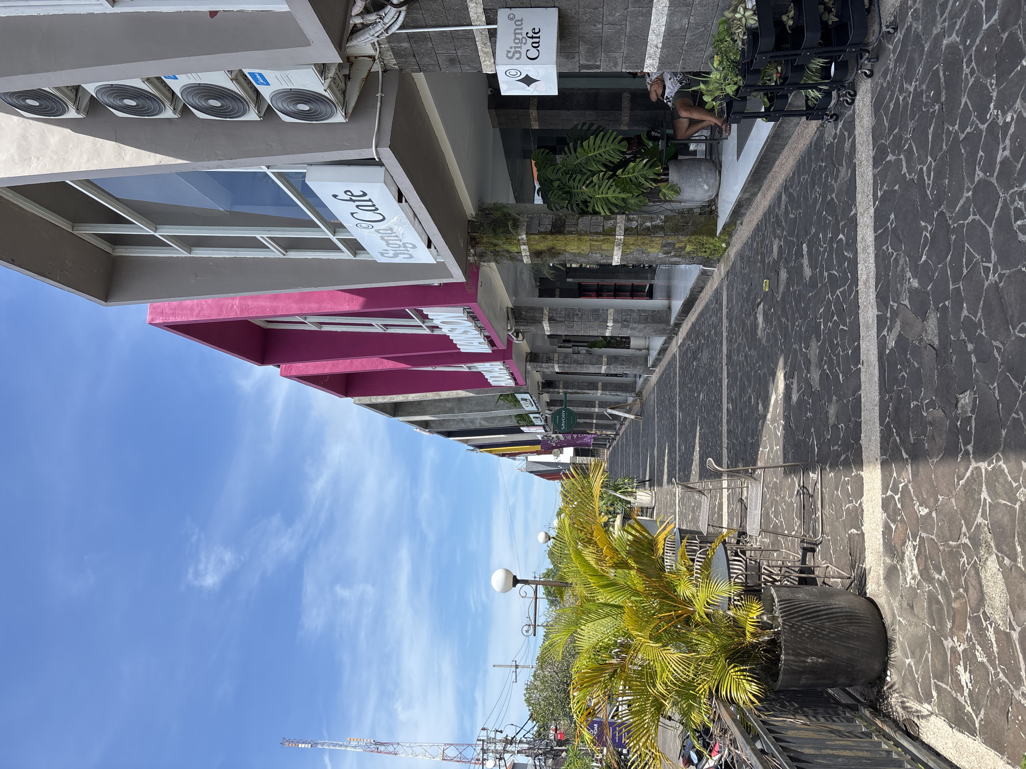 The image shows an urban street scene in Kuta Selatan, Indonesia. Buildings line the street, with signs indicating businesses such as "Signa Cafe". The buildings are varied in design and color, with one having a pink awning. Air conditioning units are visible on the facade of one building. The street is paved with dark-colored stones separated by white lines. There is an arrangement of tables and chairs along the walkway, suggesting outdoor seating for a cafe or restaurant. A large potted plant with palm-like fronds stands along the edge of the walkway. A person is seated in front of one of the buildings. In the background, there is a communication tower and power lines against a blue sky.