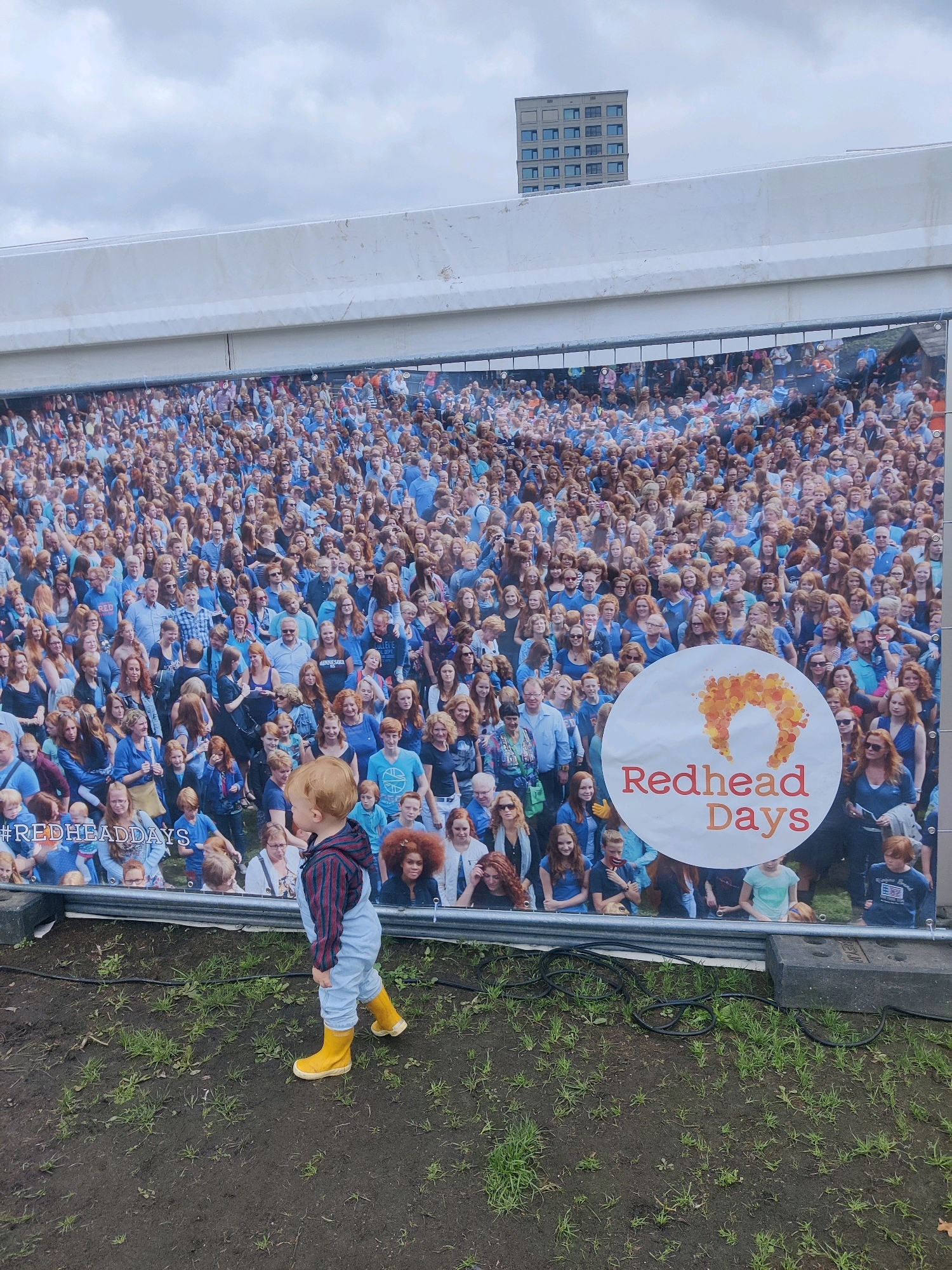 This image depicts a large banner featuring a crowd of people with red hair, promoting "Redhead Days." The banner is displayed outdoors, likely at an event or festival. In the foreground, a young child with red hair, wearing overalls and yellow boots, is walking on grass and dirt. The child is facing away from the camera, looking towards the banner. The environment appears to be a public space, with some grass and uneven ground. The sky is overcast. The banner is supported by a white structure. A tall building is visible in the background, behind the banner. The event, Redhead Days, is known to take place annually in the city of Tilburg, Netherlands.