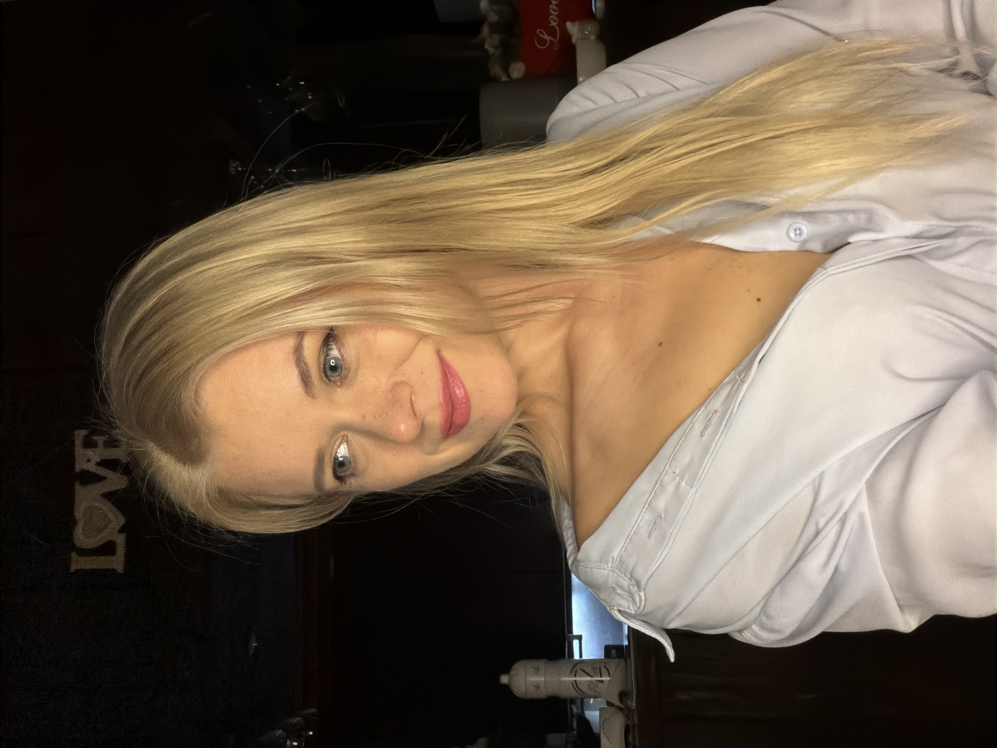 This media file is a portrait of a blonde woman wearing a light blue collared shirt. The image is shot from a close-up perspective, focusing on the woman's face and upper body. Her hair is long and blonde, cascading over her shoulders. She has blue eyes, visible makeup, and pink lipstick. The shirt is partially unbuttoned, revealing her décolletage.
In the background, a dark surface is visible, with some out-of-focus items on it. On the left side of the image, a textured object with the word "LOVE" in prominent letters is discernable. The lighting suggests an indoor setting, likely at night or in a dimly lit room, contributing to a cozy and intimate atmosphere. The woman's expression appears relaxed and her gaze is directed towards the camera. The overall mood is casual and personal. There are no obvious cues about the weather or specific location within Sochi, Russia, beyond the provided context.