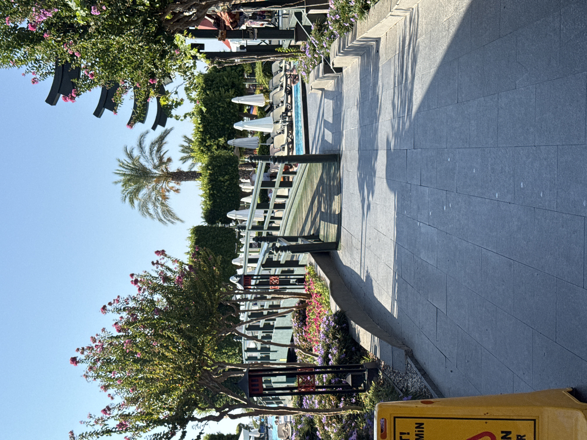 The image depicts a sunny outdoor scene at a resort or hotel in Aksu, Turkey. A pathway made of gray stone tiles leads towards a green wooden bridge with black railings. On either side of the pathway, there are trees with pink flowers and lush green bushes. A swimming pool is visible beyond the bridge, with several lounge chairs and white umbrellas. Some people are in or near the pool. In the foreground, a yellow warning sign stands on the left side of the pathway. The overall environment suggests a well-maintained recreational area.