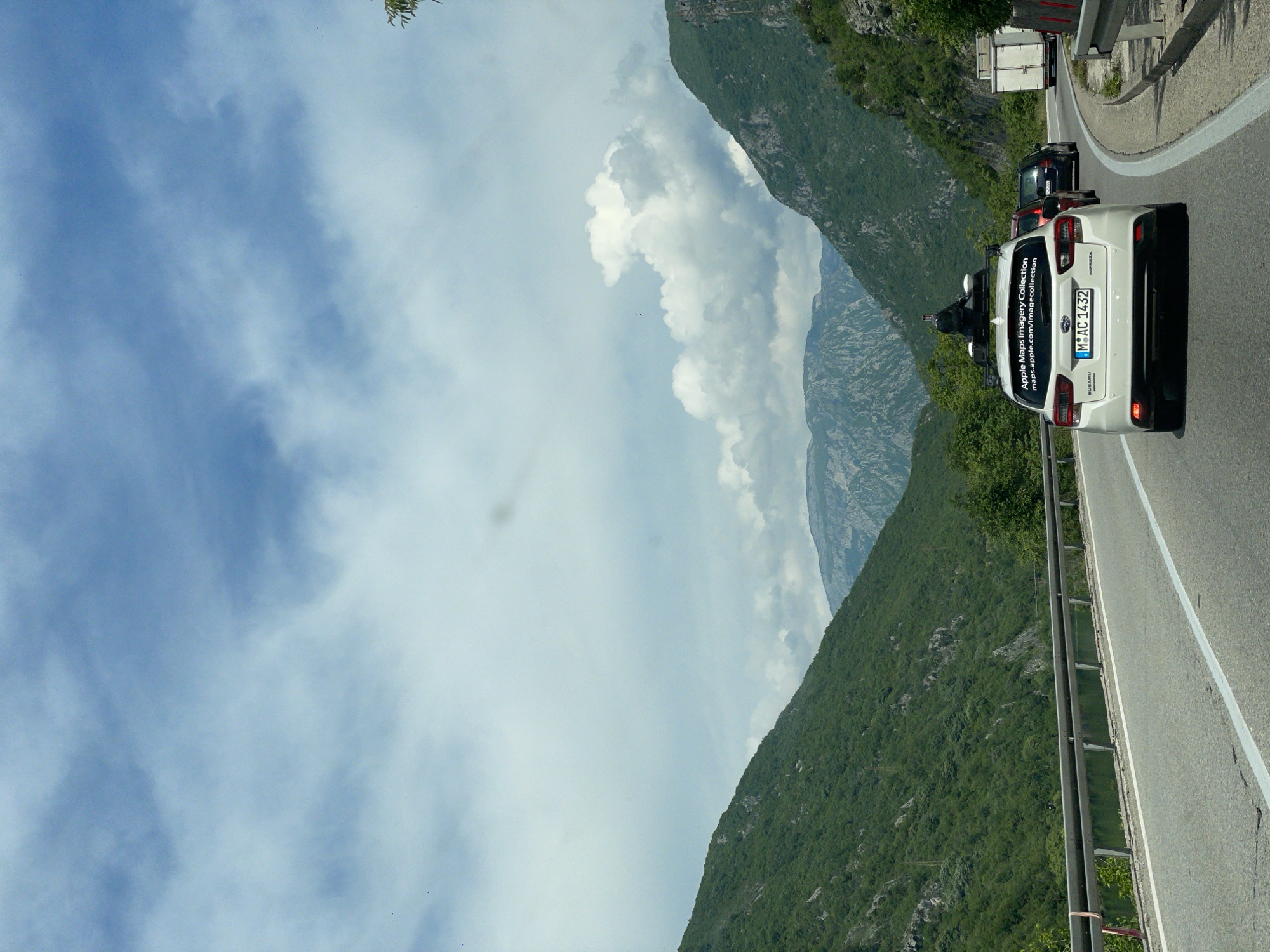 The image depicts a mountain road scene, likely in a rural area. A white car, identifiable as an Apple Maps Imagery Collection vehicle based on the sticker on the back, is driving on the road. A person is seen behind the car. A truck is visible further ahead. The road winds through a mountainous landscape, with green hills and cliffs on one side and a partly cloudy sky above. The location is potentially in Donji Jasenjani, Bosnia and Herzegovina, inferred from the Apple Maps vehicle's data collection. The perspective is from inside a car.