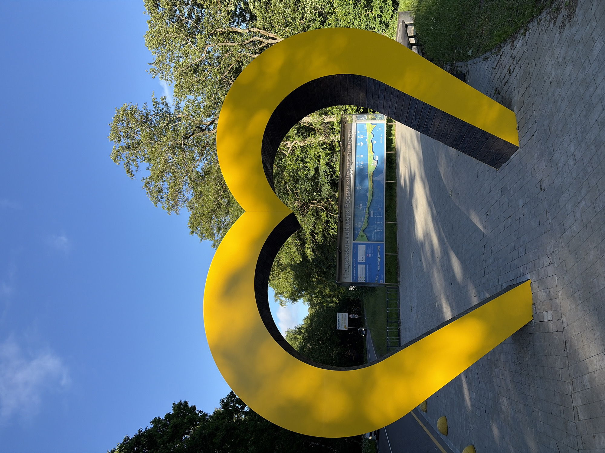 This is a vertical, eye-level, full shot of a large, bright yellow, abstract sculpture shaped like a heart. The sculpture is made of smooth metal, with a dark wood-like texture on its inner surface. It is positioned outdoors on a paved walkway, made of gray brick-like pavers. A green lawn is visible to the left of the walkway, with mature trees providing shade. A bright blue sky with a few wispy clouds is visible above, indicating a clear day. Sunlight casts shadows on the walkway, suggesting it is late afternoon or early morning.
Behind the sculpture, a large informational sign is mounted on a metal frame. The sign has a blue background and displays a map or diagram, likely related to a park or recreational area. Text is visible on the sign in a Cyrillic script. The text "НАЦИОНАЛЬНЫЙ ПАРК" is clearly legible, which translates to "NATIONAL PARK". Other text below this includes "ТУРИСТИЧЕСКИЕ ОБЪЕКТЫ" which means "TOURIST ATTRACTIONS". Further down, there is text that appears to read "РУССКИЕ РОСЫ", possibly the name of the park.
In the background, more trees line a road or path, with a few bicycles parked in a rack. The overall scene is tranquil and inviting, with the vibrant sculpture serving as a focal point. There are no people visible in the image. The environment suggests a public park or outdoor recreation area.