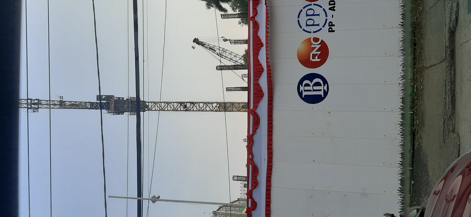 The image shows a construction site in Jambi, Indonesia. A large tower crane is visible against a clear sky, along with another smaller crane. In the foreground, a white hoarding with decorative red and white draped fabric at the top displays several logos. These logos include a blue circle with a white "B" inside, an orange circle with "FNC" in white lettering, and a blue circle with "PP" inside, accompanied by the text "PP - AD". Below the logos, a stylized green grass design is painted along the bottom of the hoarding. The scene is viewed from a street level, with a portion of a red vehicle in the lower left corner and a damaged road surface in the foreground. Overhead power lines crisscross the upper portion of the image.