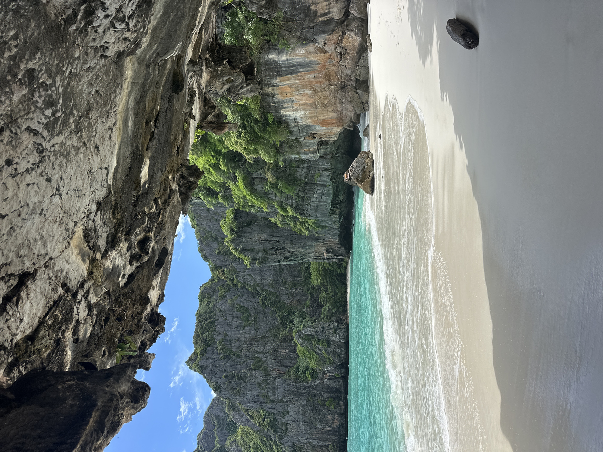 The image captures a serene beach scene in Ao Nang, Thailand, framed by the overhang of a cave. The foreground is dominated by the textured, rough surface of the cave ceiling, casting a shadow onto the pristine white sand below. The sand slopes gently towards the turquoise waters of the Andaman Sea, where gentle waves create foamy white crests as they reach the shore.
In the midground, lush green vegetation carpets the towering limestone cliffs that rise dramatically behind the beach. These cliffs, characteristic of the region, are rugged and weathered, with patches of exposed rock showing warmer, rust-colored hues. A few large boulders are scattered on the beach, one of which is large enough to have a lone figure sitting atop it, observing the tranquil vista.
The sky above is a clear, bright blue, with a few wispy white clouds. The overall impression is one of natural beauty and peaceful isolation, with the cave providing a natural frame to the stunning coastal landscape. The scene is devoid of any man-made structures, emphasizing the untouched nature of this tropical paradise. The lighting suggests it is daytime, likely late morning or early afternoon, given the strong shadows.