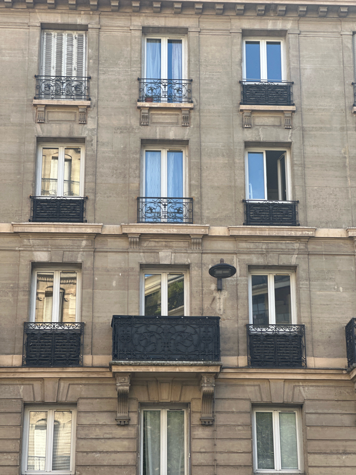The image depicts the facade of a multi-story building in Paris, France. The building has a neoclassical architectural style, characterized by its stone construction, symmetrical window placement, and ornate decorative elements. The facade is divided into multiple floors, each featuring several windows and balconies.
The windows are predominantly white-framed and appear to be double-paned. Some windows are covered with white shutters, while others reveal glimpses of blue curtains. Several windows have small, black wrought-iron balconies with decorative railings. One balcony on the second level from the top features an open window above it, suggesting a door or entrance.
The building's exterior is constructed of light brown stone blocks, with horizontal banding and vertical pilasters separating the window sections on some floors. Decorative stone moldings are present above and below the window levels. On the third level from the top, a black, circular light fixture is mounted to the wall between two windows.
The lower section of the building shows more windows with white frames and blinds. The windows reflect the surroundings, including other buildings and possibly trees. The overall impression is one of a well-maintained, historic Parisian building. There are no people visible in the image.