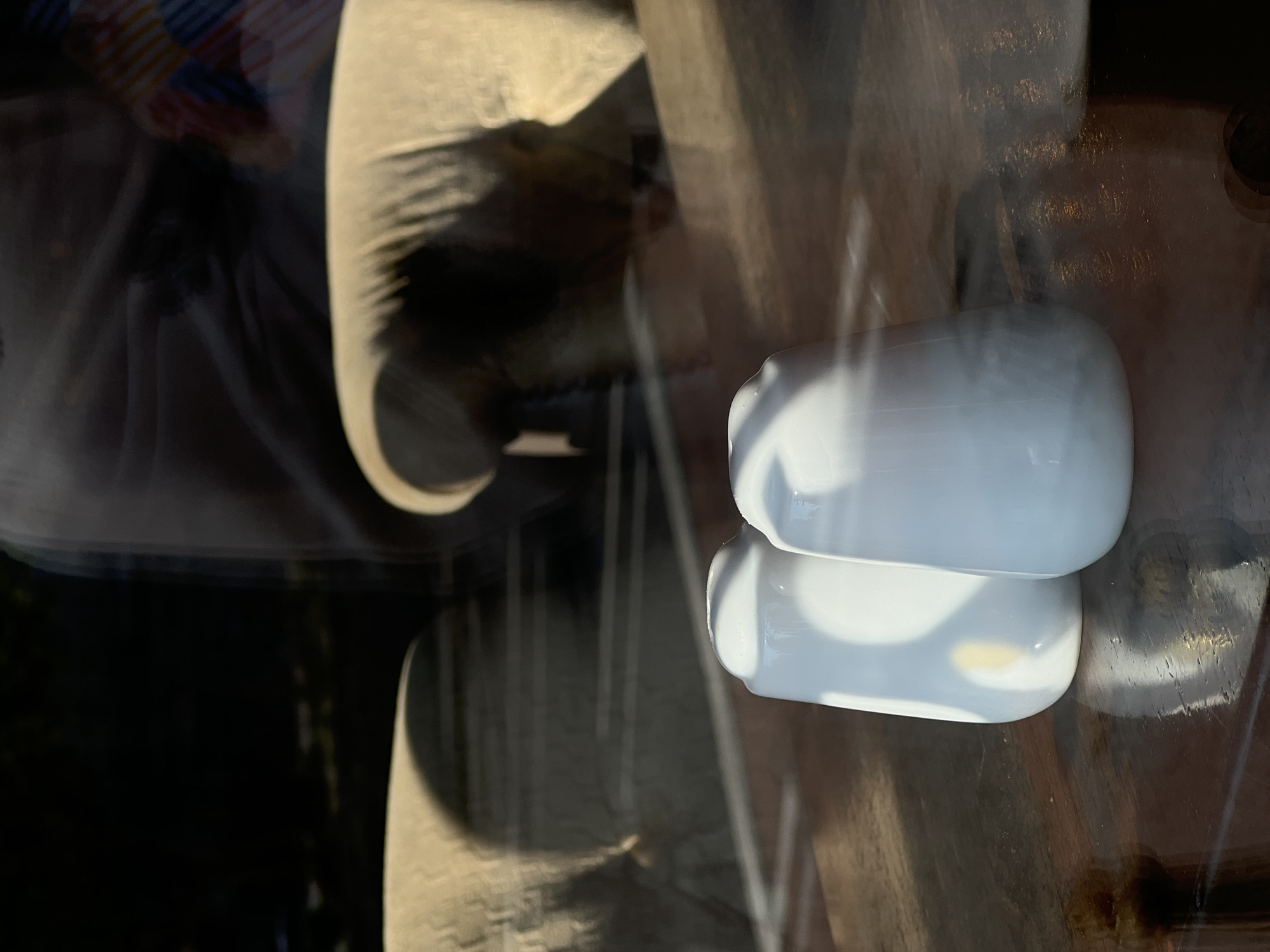 The image shows two white ceramic salt and pepper shakers on a dark wooden surface. The shakers are conical in shape with rounded tops. They are positioned close together, with the one on the left slightly behind and to the left of the one on the right. Sunlight casts strong shadows on the wooden surface and illuminates the shakers. In the background, through what appears to be a window, there are blurred reflections of furniture and a person. The overall scene suggests an indoor setting, possibly a restaurant or dining area.
The location is identified as the city of Donegal, Ireland. This detail is provided without any visual cues in the image itself to support it.