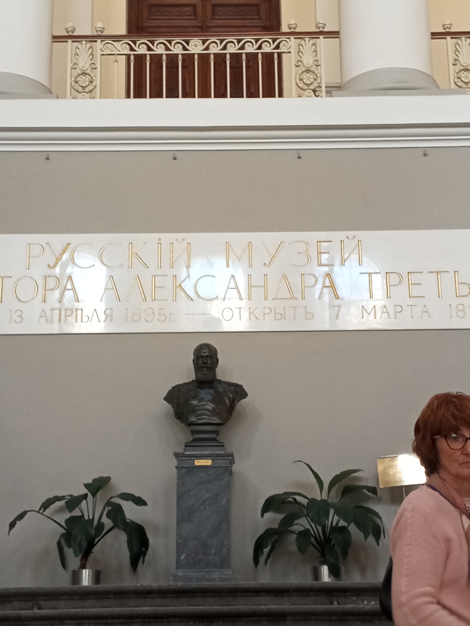 The image depicts the interior of the Russian Museum (Русский музей) in Saint Petersburg, Russia.
A plaque mounted on a wall displays the inscription "РУССКІЙ МУЗЕЙ ТОРА АЛЕКСАНДРА ТРЕТЬ 13 АПРГЪЛЯ 1895г. - ОТКРЫТЬ 7 МАРТА 18:".
Below the plaque is a dark bust of a man, placed on a gray pedestal. On the horizontal surface below the bust and plaque are two potted plants.
To the right, a woman with red hair and glasses is partially visible. The background shows architectural details of the interior, including a railing and columns.