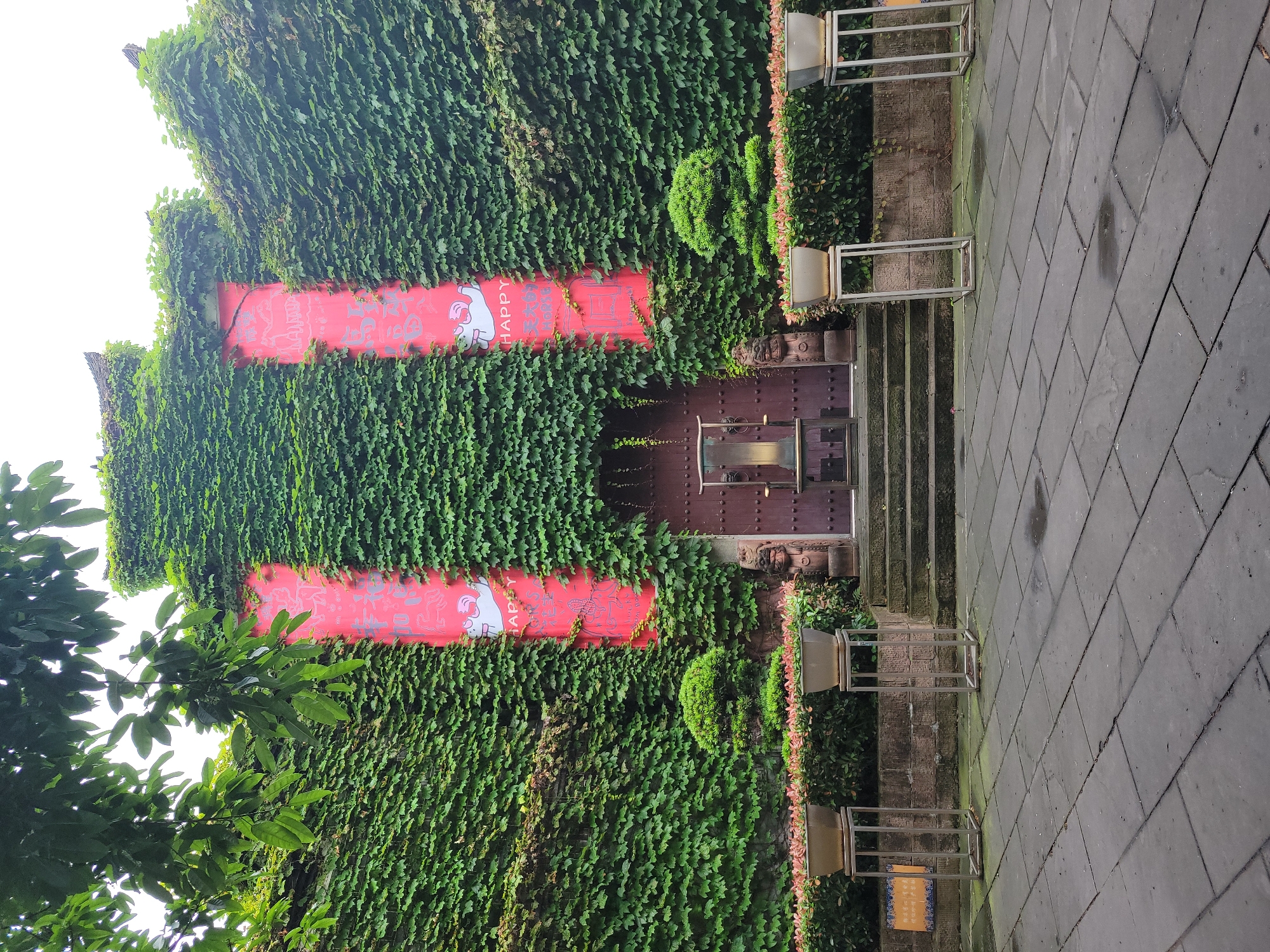 The image shows a building facade in Hechuan, China, completely covered in dense ivy. The entrance is centered with a dark wood door reached by a few stone steps. Flanking the door are two vertical red banners with the word "HAPPY" in English and what appears to be Chinese characters and a stylized graphic. Metal frames containing planters sit on either side of the steps. The ground is paved with large gray stone tiles. Tree branches are visible in the lower left corner.