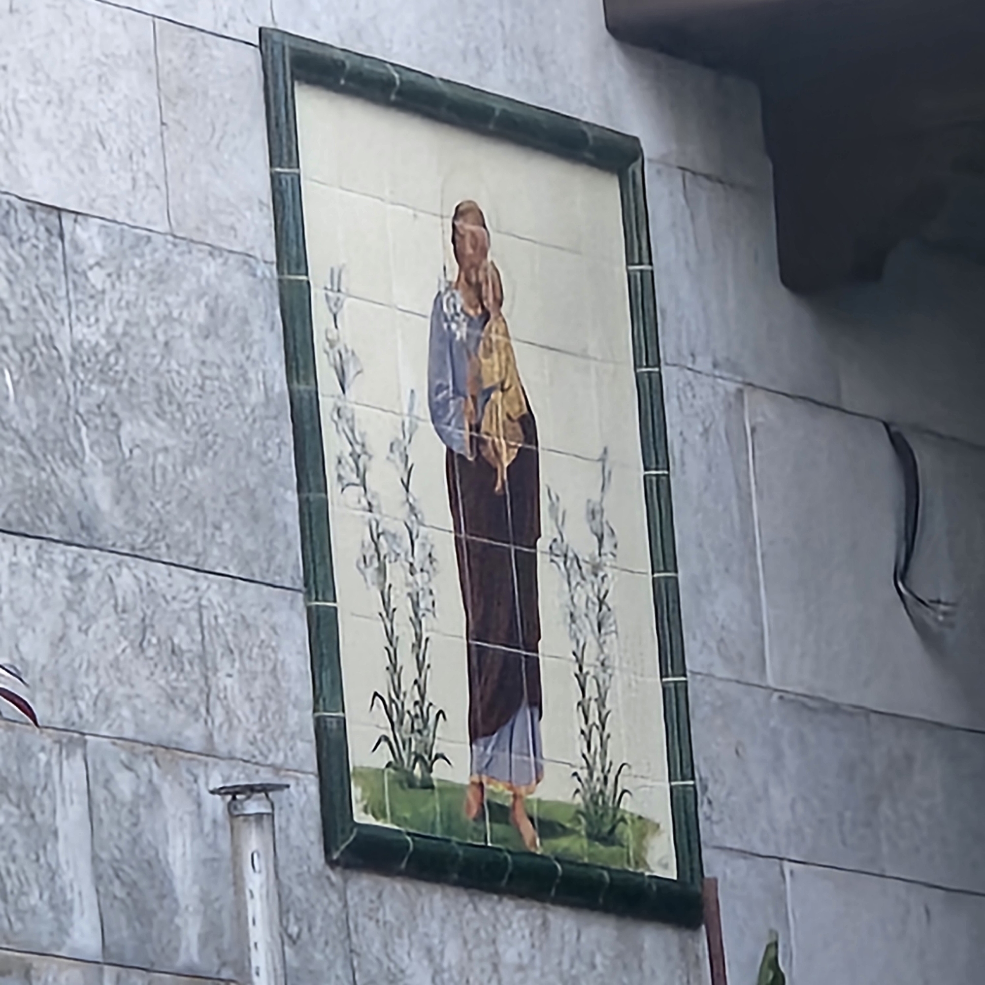 An outdoor ceramic tile mural, framed by a dark green border, depicts a religious figure in a dark robe holding a child wrapped in a gold cloth. The figure has a halo and stands amidst stylized white flowers. The mural is mounted on a light gray stone wall, possibly in the city of Aiguaviva, Spain, judging by the architectural style and common practice of such art in the region. To the left of the mural, a portion of a white vertical pipe with horizontal lines is visible. The lighting suggests it is daytime.