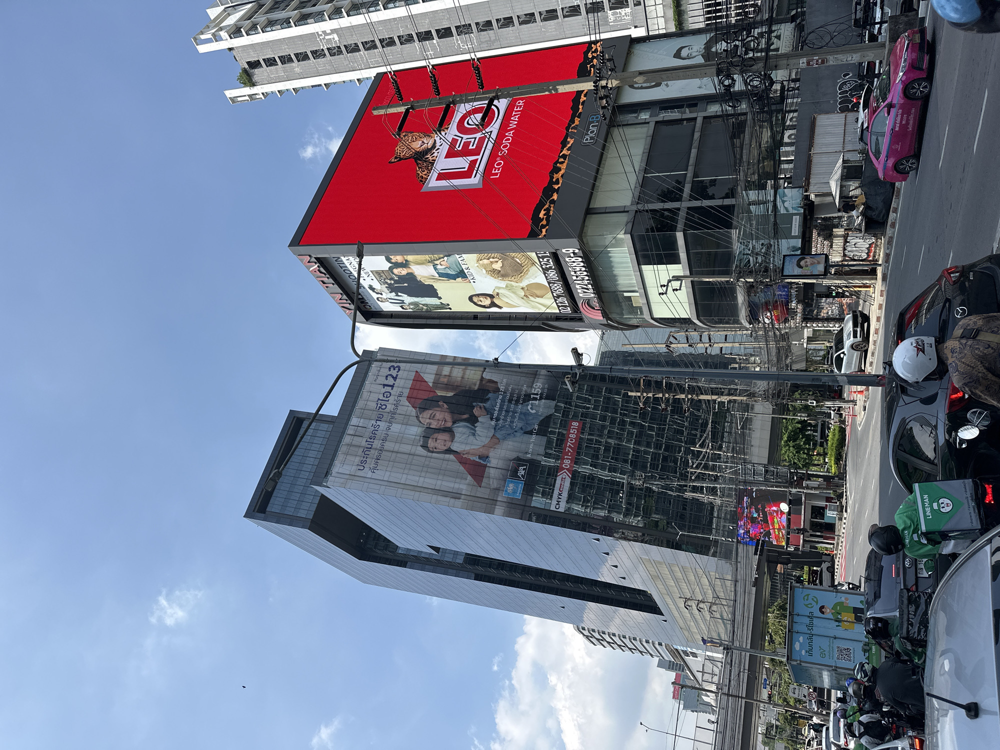The image captures a bustling street scene in Bangkok, Thailand. The view is oriented vertically, focusing on the dense urban landscape. Tall buildings dominate the backdrop, including modern structures with reflective glass facades and large advertising billboards. The most prominent billboard features a red background and advertises "LEO Soda Water" alongside other advertisements for products such as CMYK Credit insurance and AXA insurance. The streets are crowded with traffic, including cars and a large number of motorcycles, some with delivery services branding like "LINE MAN". Power lines and utility wires form a complex web in the foreground, obscuring parts of the buildings and sky. The sky is partly cloudy with a clear blue color. The image depicts a vibrant and busy urban environment characteristic of Bangkok.