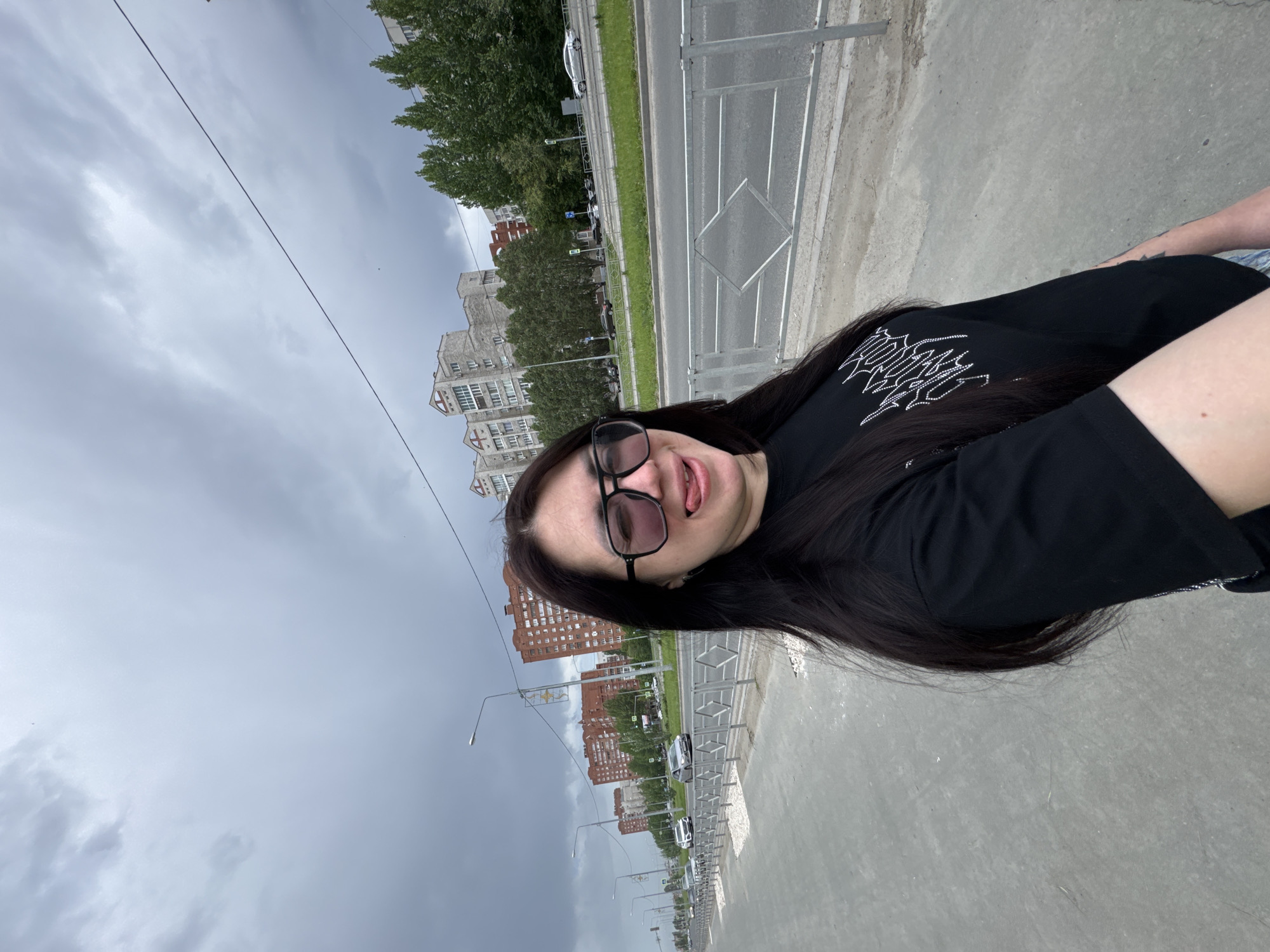A woman with long, dark hair, wearing black-framed glasses and a black t-shirt, is standing on a concrete sidewalk next to a road. She is sticking her tongue out and looking at the camera. The road has a metal guardrail and a grassy median. Beyond the road are rows of apartment buildings, with trees interspersed. The sky is overcast. The location is in a city, likely in Unknown, Unknown, indicated by the urban infrastructure and apartment buildings.
