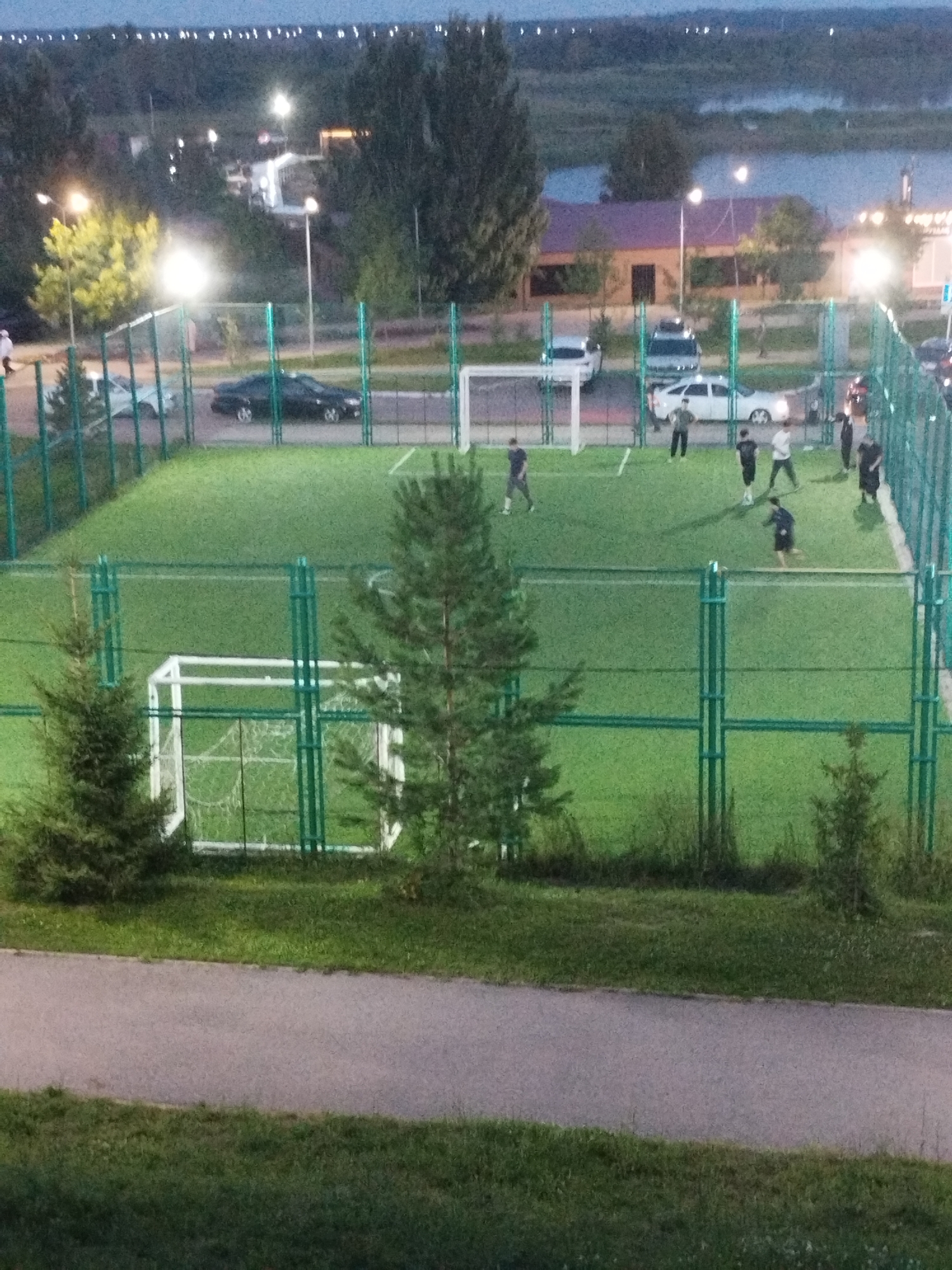 The image depicts an outdoor artificial turf soccer field at dusk, likely in Kostanay, Kazakhstan. The field is enclosed by green metal fencing. Several individuals, appearing to be young men, are engaged in a soccer game. One person is near a white soccer goal on the left side of the frame, while others are scattered across the field, some actively participating in the game. The surrounding environment shows trees, grass, a paved pathway, and in the background, buildings, streetlights, and a body of water, possibly a river or lake, are visible. The sky is dark with some lights from distant structures. Cars are parked on a road adjacent to the field.