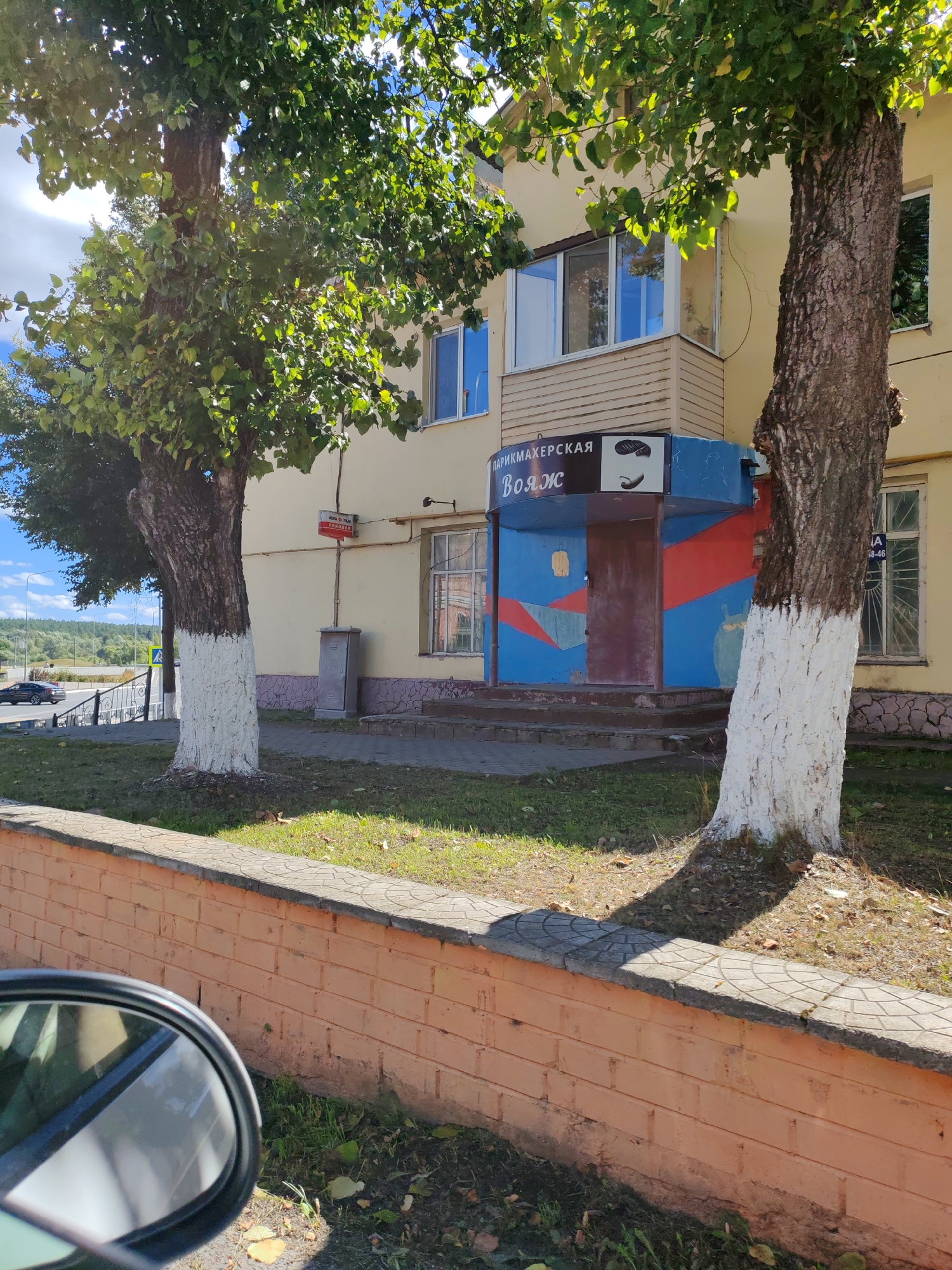 The image shows the facade of a building in the city of Kirov, Russia. A bright yellow, two-story building is visible, with trees in the foreground. One tree, on the right side of the image, has its trunk painted white at the base. The building has several windows, some with balconies. A blue and red painted entrance area, possibly for a business, is located between two trees. Text above this entrance reads "ПАРИКМАХЕРСКАЯ" (hairdresser's) and "ВОЯЖ" (voyage). A red sign with white lettering, possibly a company logo, is mounted on the wall of the building. The area in front of the building is paved with sidewalk tiles and has patches of grass and fallen leaves. A low orange brick wall with a decorative concrete border runs along the front of the scene. In the background, a road with a passing car and a fence are visible, along with a wooded area and a cloudy sky. The overall scene suggests a commercial or residential area in a suburban or urban setting.