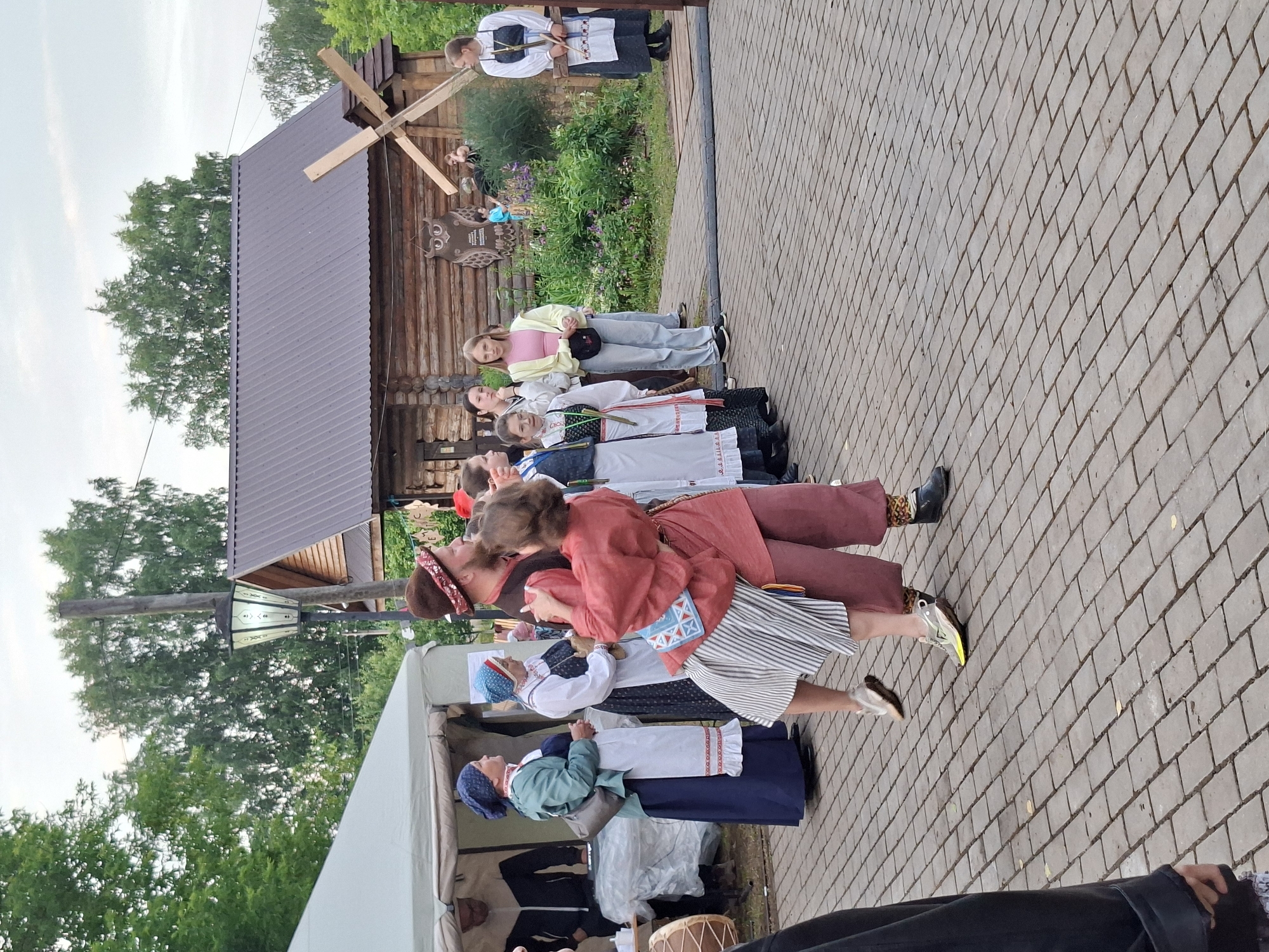 In the city of Старые Быги, Russia, a group of people are gathered in what appears to be an outdoor cultural event or festival. The foreground features a couple in traditional attire dancing, with the woman wearing a striped skirt and the man in a maroon outfit. Behind them, several women in traditional Russian clothing are observing, some standing by a tent that displays various items. To the right, a wooden building with a pitched roof stands prominently, adorned with decorative elements like an owl carving and a windmill. Other individuals in a mix of modern and traditional clothing are also present, observing the festivities or engaged in other activities. The ground is paved with cobblestones, and trees and greenery surround the area, suggesting a rural or historical setting.