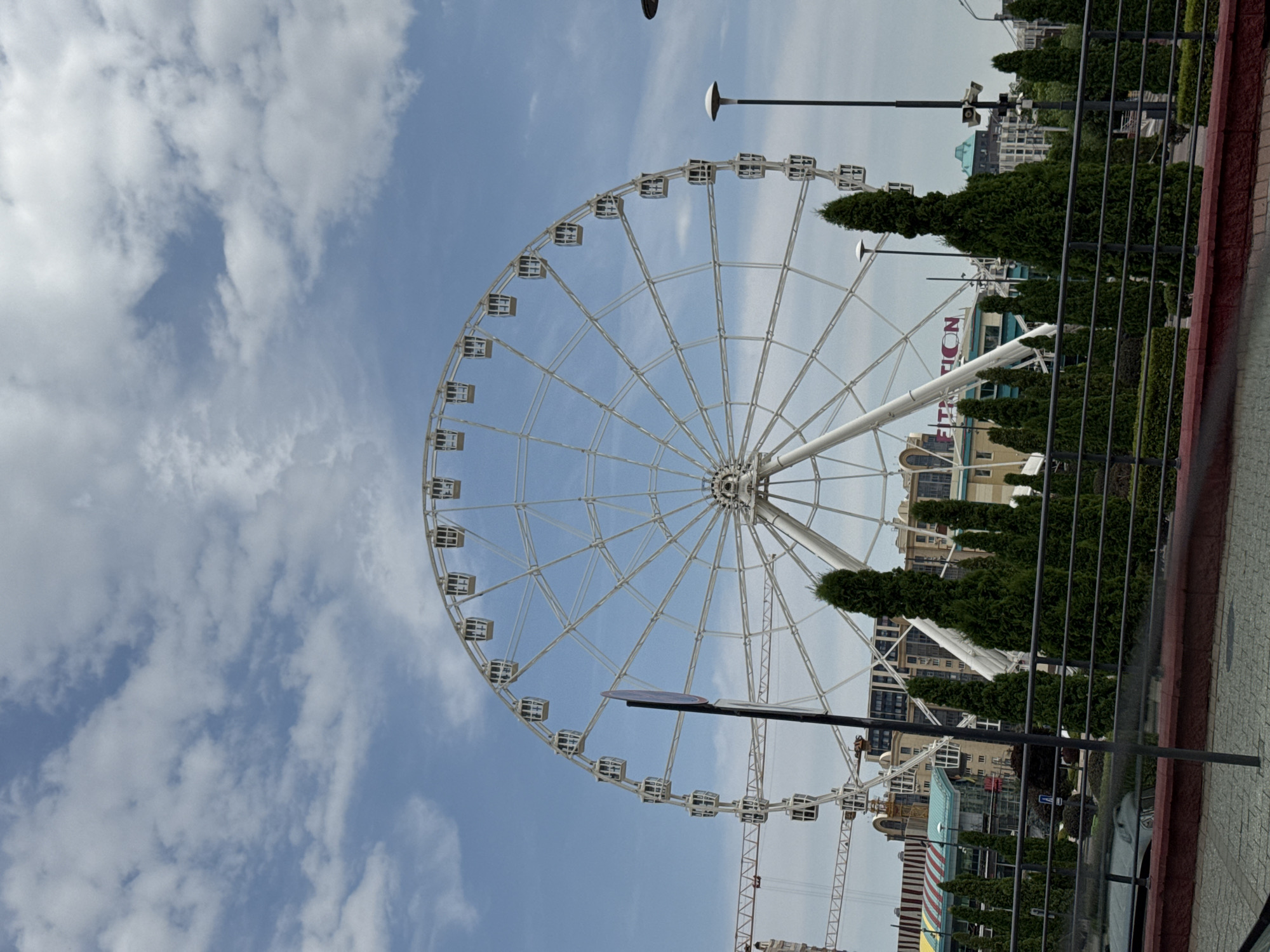 An outdoor, daytime, eye-level, vertical shot shows a large Ferris wheel in the city of Almaty, Kazakhstan. The Ferris wheel is white and has many gondolas. In the background, there are several buildings, including one with a sign that appears to say "FITNESS." There are also trees and a cloudy blue sky. The foreground shows a walkway and a fence.