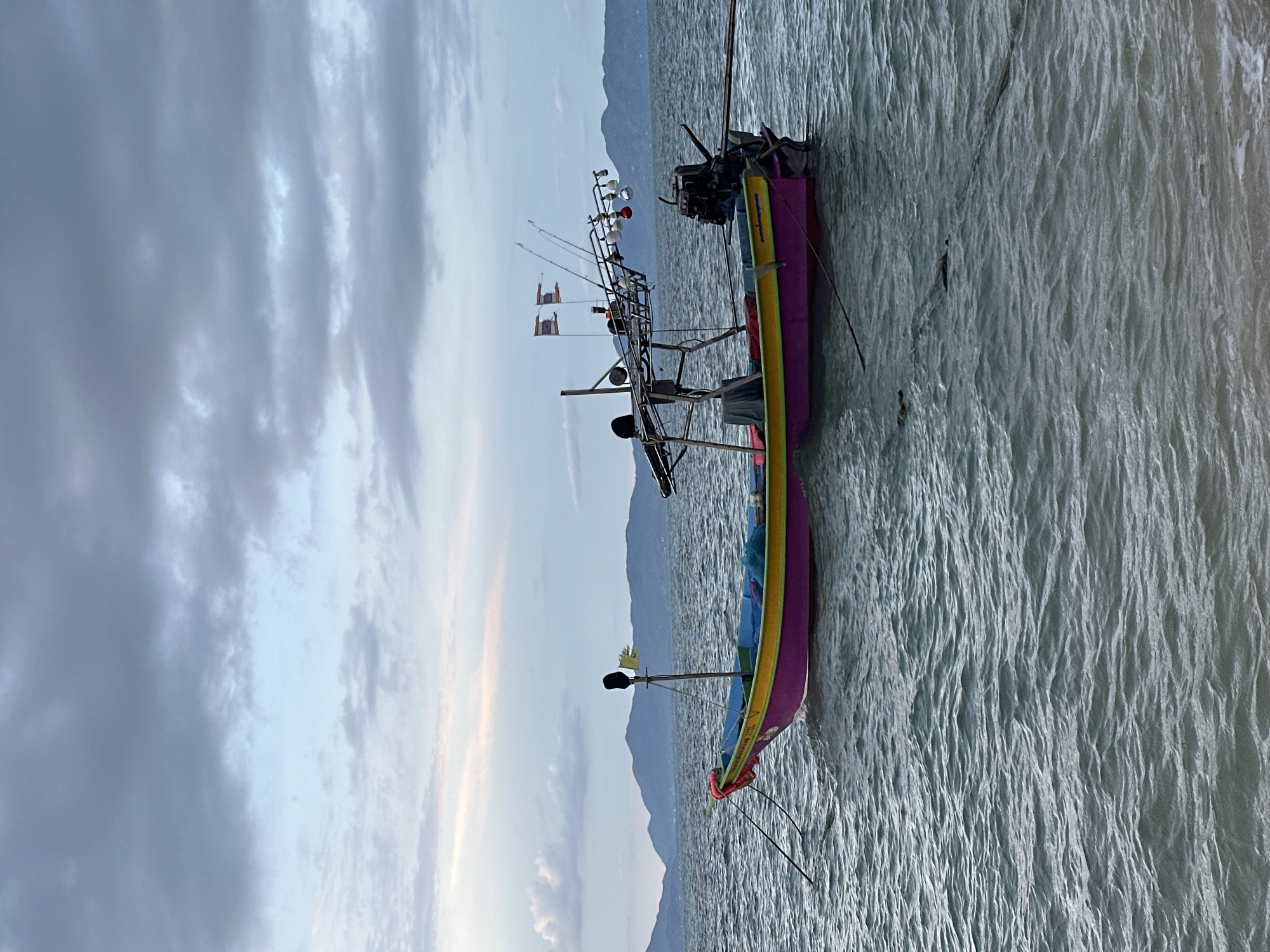 This image, taken in Baan Thong Po, Thailand, depicts a tranquil scene of two fishing boats moored in the water. The boats are colorful, with the closest one featuring vibrant purple, yellow, and blue hues. It is equipped with fishing rods, multiple lights, and a motor at the stern. A small flag is also visible on the boat's rigging. The second boat, partially obscured by the first, shares a similar structure and appears to be anchored.
The boats are situated on a body of water with a choppy surface, indicating a moderate breeze. In the background, a hazy mountain range stretches across the horizon, suggesting a coastal or island location. The sky is overcast with a mix of dark gray and lighter, softer clouds, hinting at either early morning or late afternoon. The overall atmosphere is peaceful and serene, capturing a moment of quiet before or after a fishing excursion. No people are visible in the image, emphasizing the solitude of the scene. There is no visible text in the image.
